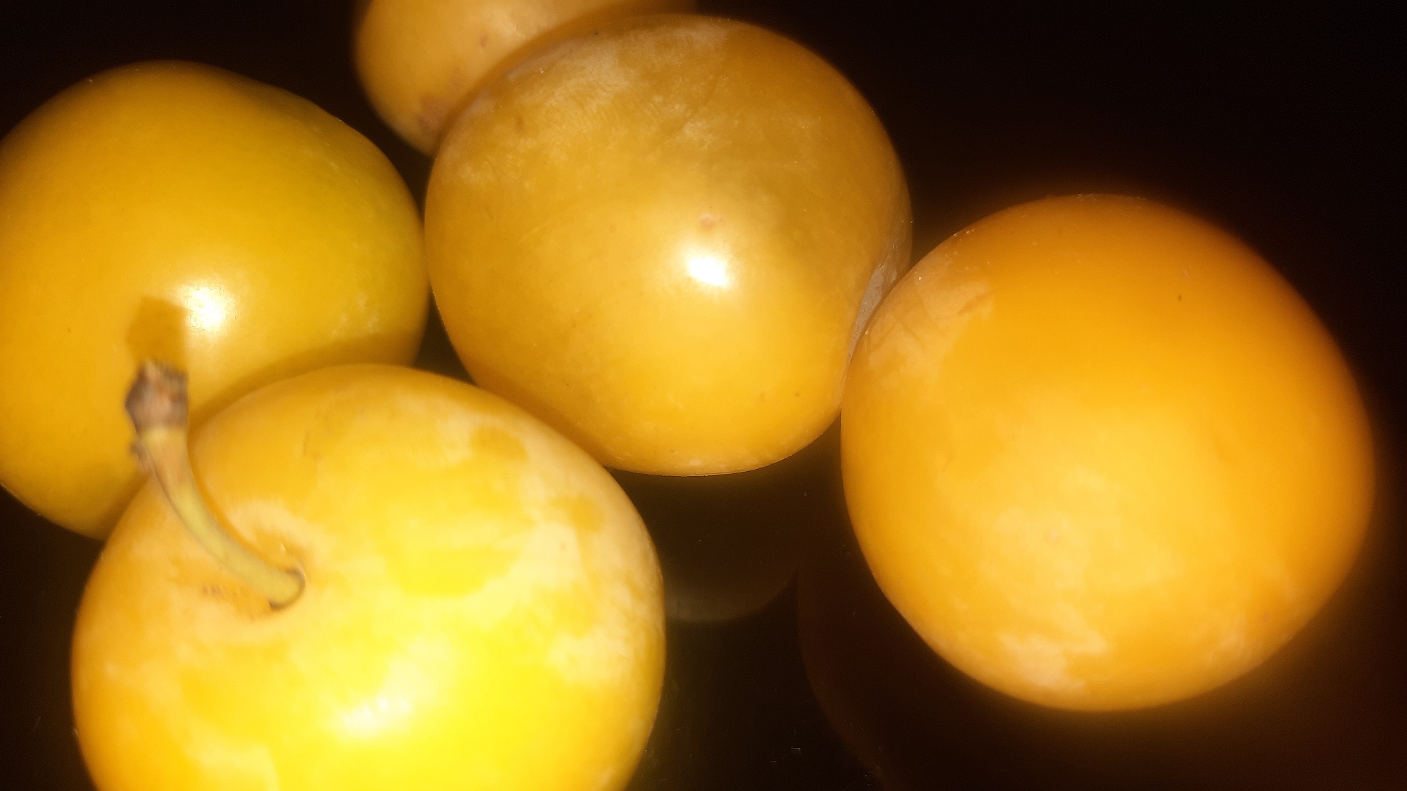 A close-up, overhead shot shows five yellow plums arranged in a cluster on a dark, reflective surface. The plums are round and smooth, with a slightly matte finish that has a subtle sheen in places. Some of the plums are positioned on top of others, creating layers within the arrangement. The lighting is dim, highlighting the smooth surfaces and contours of the fruit. The background is entirely black, focusing all attention on the plums. There is no visible indication of the location being in the city of Шклоў, Belarus.