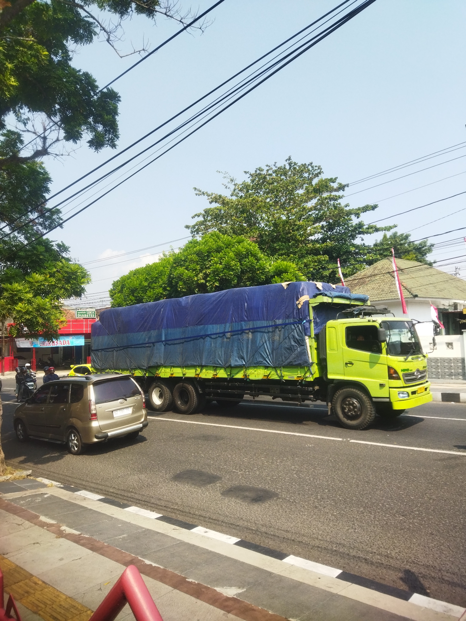 A bright green truck with a blue tarp-covered cargo bed is driving on a paved road in Giripurwo, Indonesia. The truck is the main subject of the image and is positioned in the center. To the left of the truck, a gold-colored van is parked on the side of the road. In the background, there are several buildings and lush green trees. The sky is clear and blue. Power lines are visible crisscrossing the sky. There are also some motorcycles parked near the buildings.