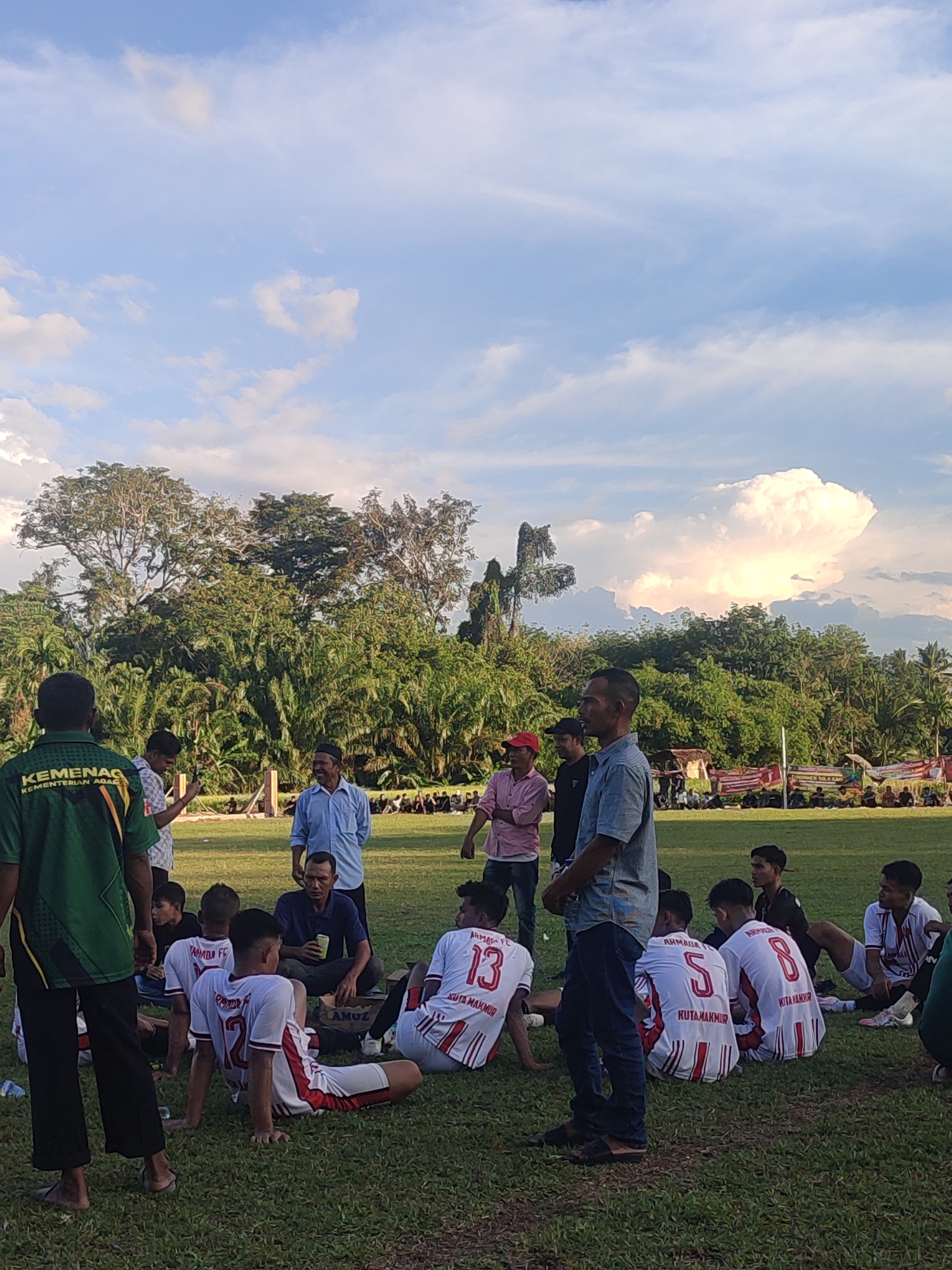 A group of young men, identifiable as soccer players by their uniforms, are gathered on a grassy field under a partly cloudy sky. Most of them are sitting, forming a semi-circle, while a few adults stand around them, engaging in conversation. The players' jerseys are predominantly white with red stripes, each displaying a number and the words "KUTA MAKMUR". In the background, there's a line of trees and what appears to be a banner or decoration. The scene suggests a break or debriefing during a soccer event. The location is presumed to be in Babahlueng, Indonesia.