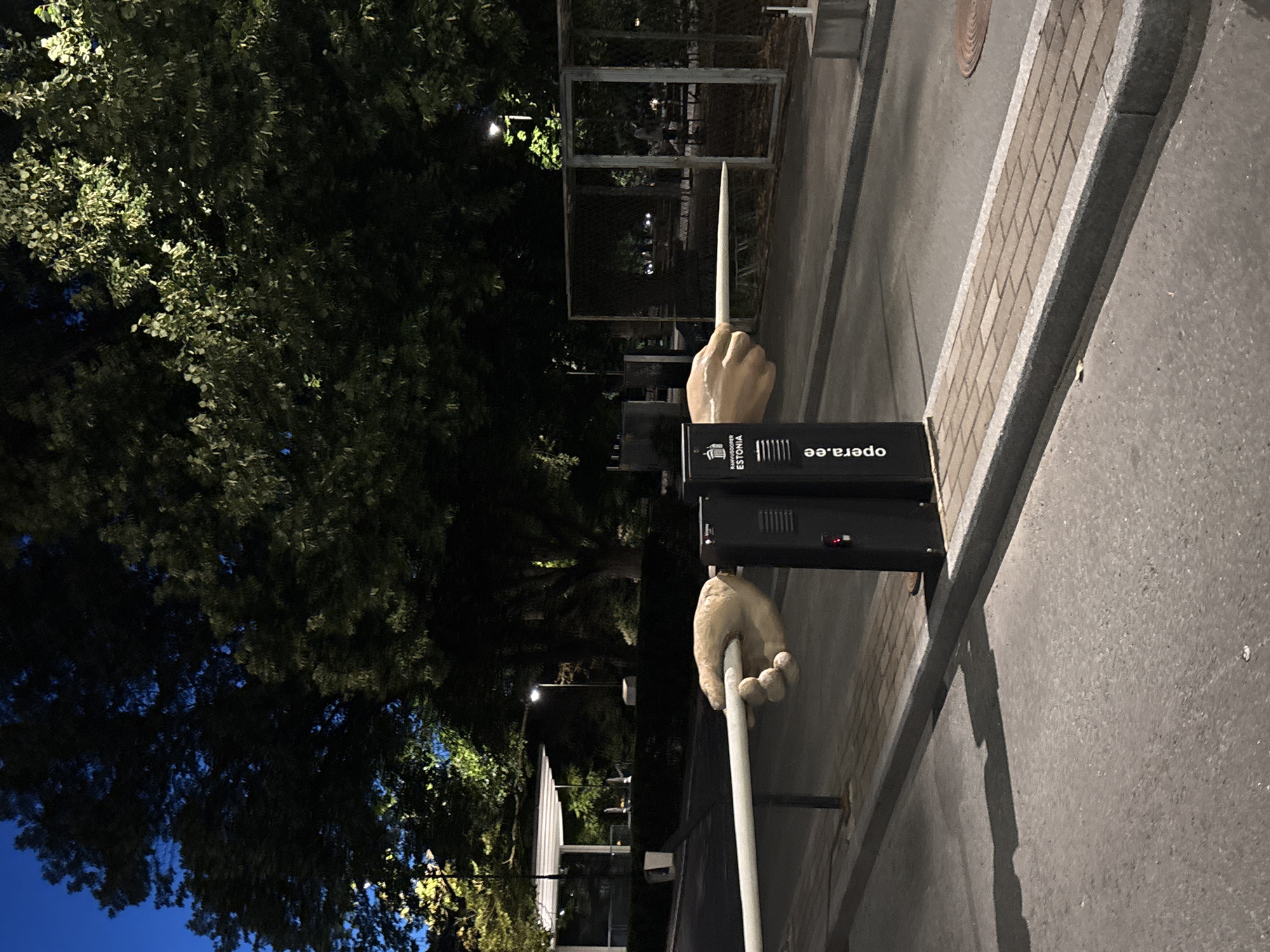 The image captures an outdoor scene at night in Tallinn, Estonia, near the opera house, indicated by the "opera.ee" text on a black pillar. The foreground is dominated by an asphalt road and a paved walkway. A white barrier pole is prominently displayed, seemingly held in place by two large, realistic sculptures of hands. One hand grips the pole from the left, and another from the right. These hands appear to be part of a sculptural installation.
To the right of the hands, a black electronic pillar stands, also featuring the "opera.ee" text and an Estonian national emblem. Behind the barrier and hands, there is a paved area leading to a fenced enclosure with benches and trees. Further in the background, large trees with illuminated leaves suggest artificial lighting. A white canopy structure is visible in the far left background, possibly part of a building. The sky is dark blue, indicating nighttime. The overall environment is urban, likely a public space associated with the opera house. There are no visible people in the immediate vicinity of the sculptural hands or barrier.