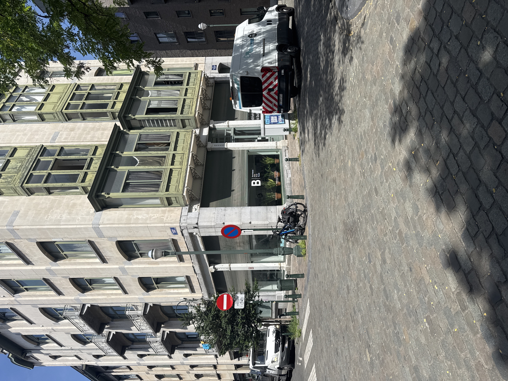 In a city intersection with a cobblestone street, a white van with reflective chevron markings on its rear is parked on the right. Adjacent to the van, a blue bicycle is parked near a lamppost. On the left side of the image, a white delivery van is parked in front of a building with balconies. Further down the street, a tree stands next to a building with closed storefronts and a sign with the letter "B" prominently displayed. Two traffic signs are visible: a red circle with a white horizontal bar on the left, and a blue circle with a red diagonal bar on the right. The buildings in the background are multi-story structures, with some featuring green-trimmed bay windows. The sky is clear and blue.