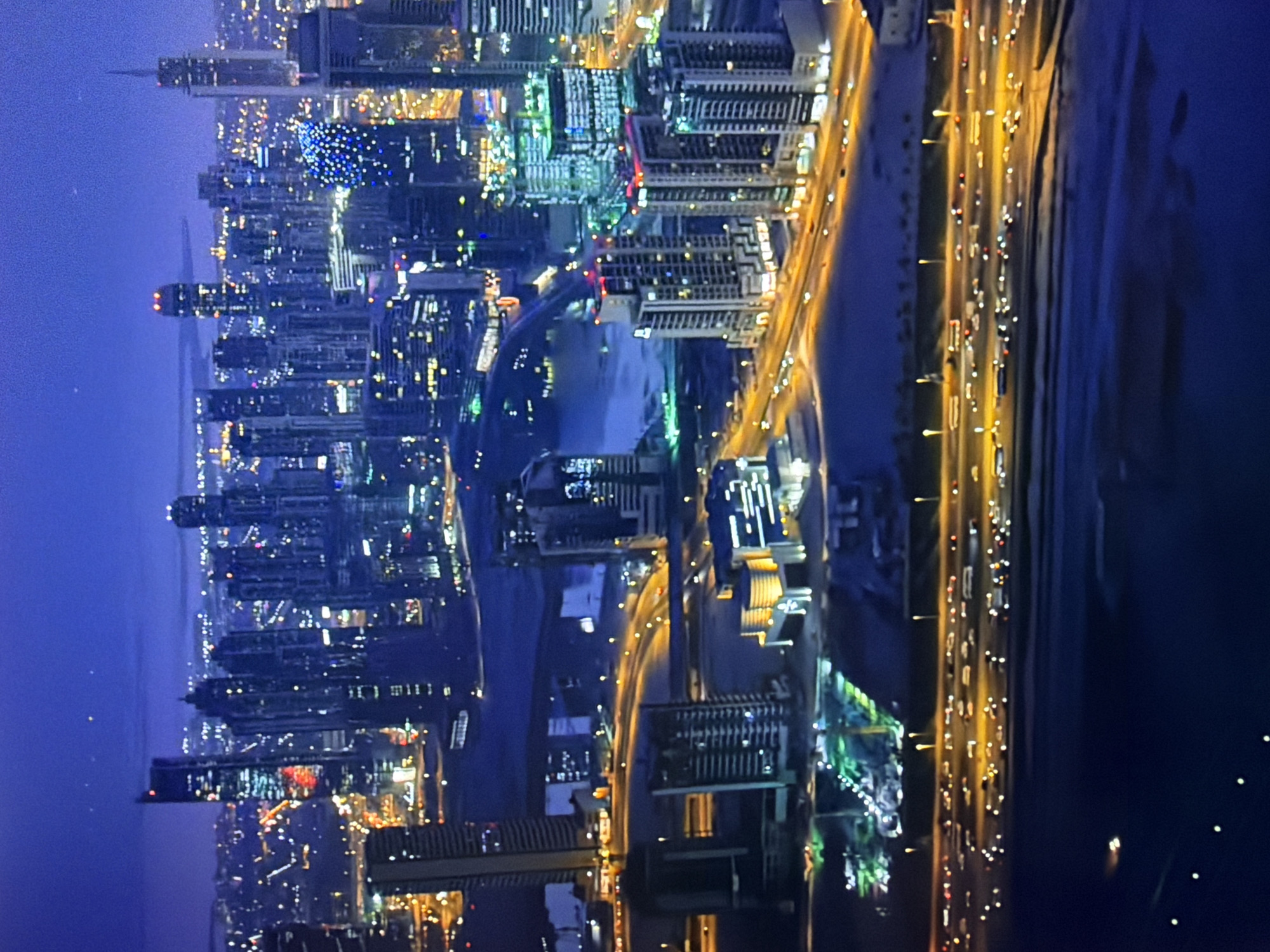 This is a nighttime aerial view of a dense urban landscape dominated by modern skyscrapers. The buildings are illuminated with numerous lights, creating a vibrant, electric atmosphere. A network of roads with flowing traffic, indicated by streaks of light from headlights and taillights, crisscrosses the scene. The overall impression is one of a bustling metropolis at night, likely a major city due to the scale and modernity of the architecture. The specific location is not identifiable as Kyiv, Ukraine, based solely on the visual information presented in the image.