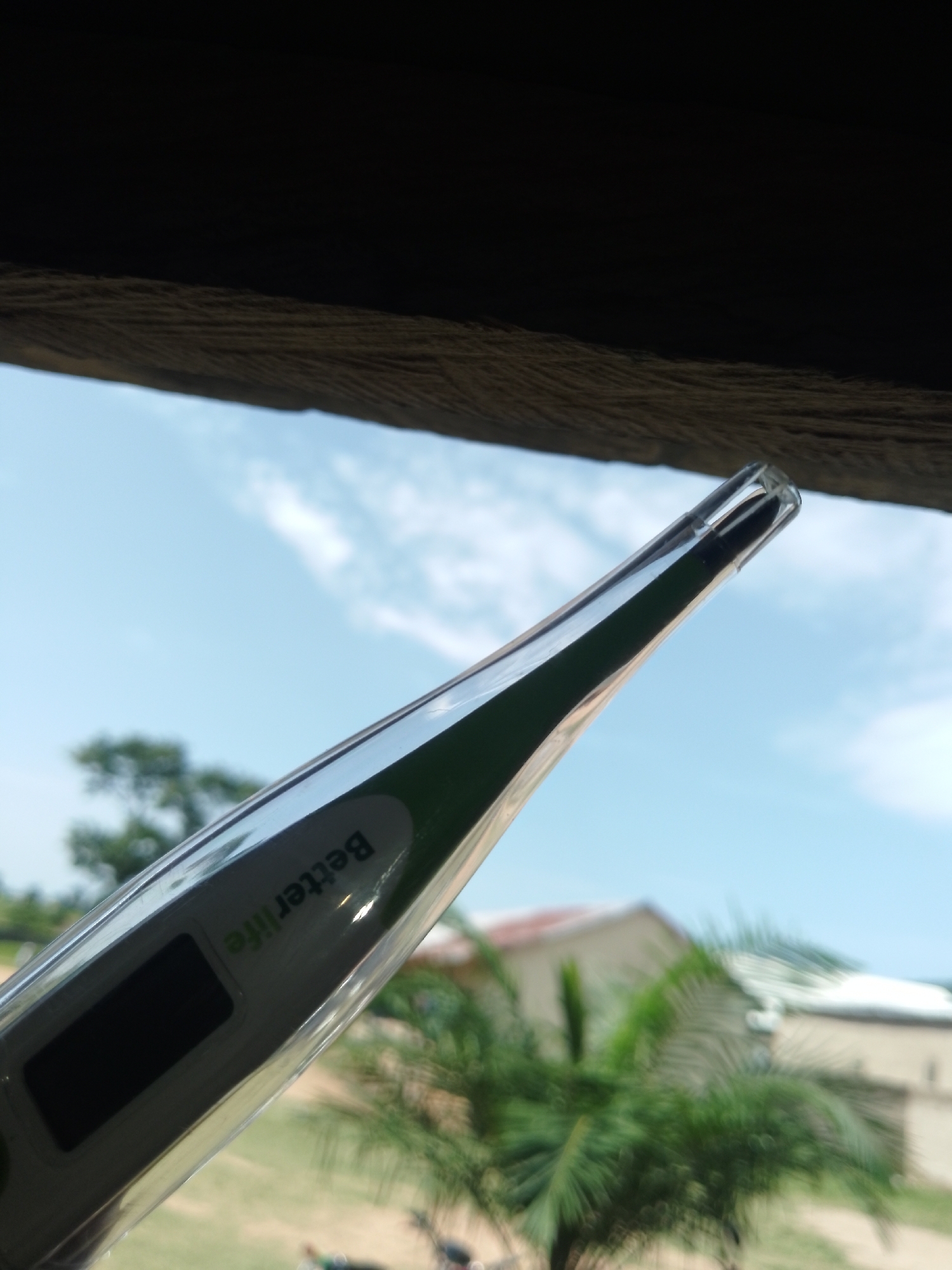 A digital thermometer with "BetterLife" branding is held up against a blue sky with scattered white clouds, seen from below. The thermometer is angled upwards, with its clear plastic tip extending to the right. A dark, rectangular display is visible on the side of the thermometer. In the background, out of focus, are green trees, palm fronds, and parts of buildings with red and white roofs. The top of the image is dominated by a dark, textured surface, possibly a wooden overhang. The overall scene suggests an outdoor setting.