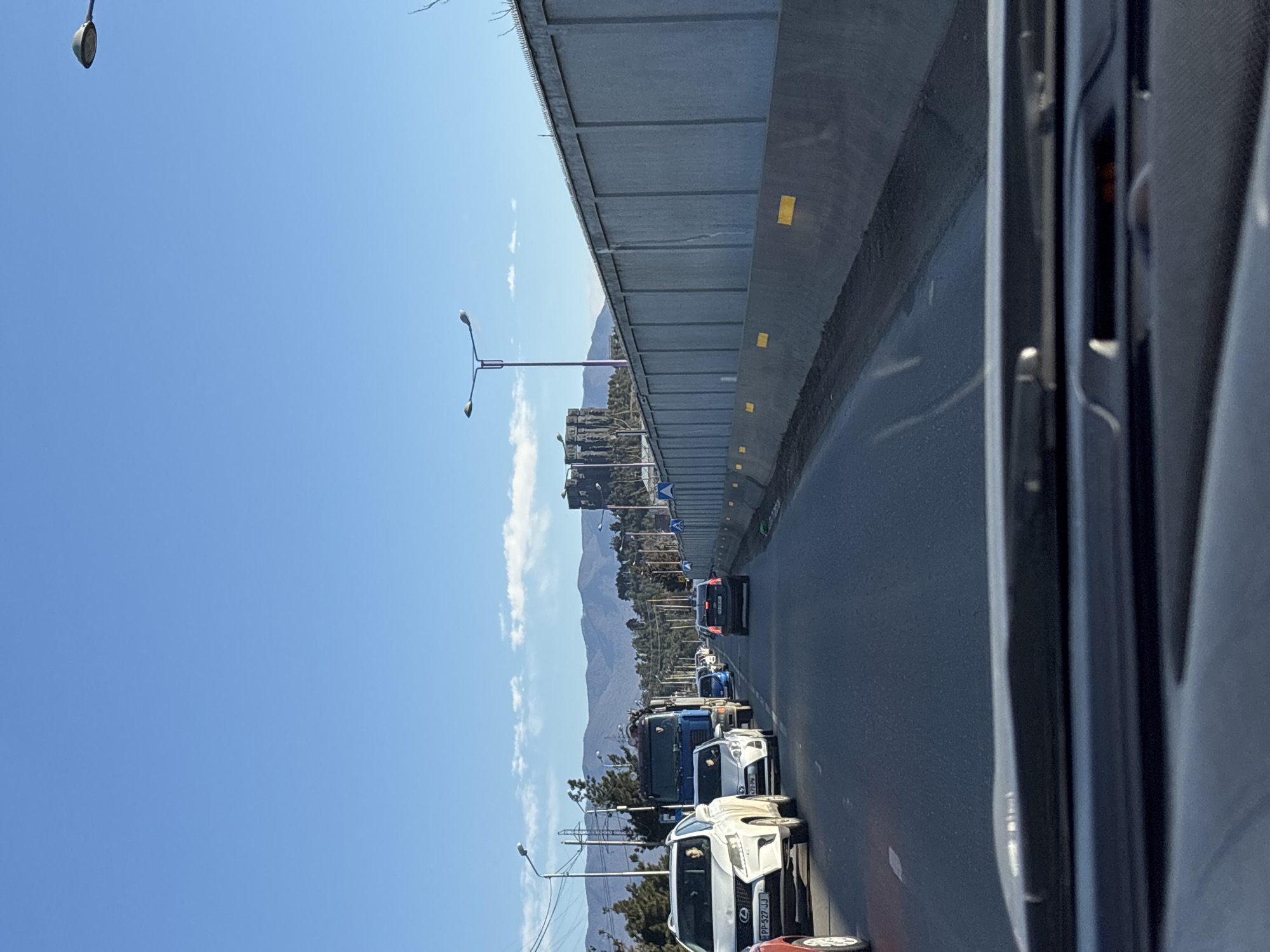 The image shows traffic on a road in Tbilisi, Georgia. The road is under an elevated highway or overpass. In the distance, there are mountains. Cars and a truck are visible in the traffic. The sky is blue. The view is from inside a vehicle.
