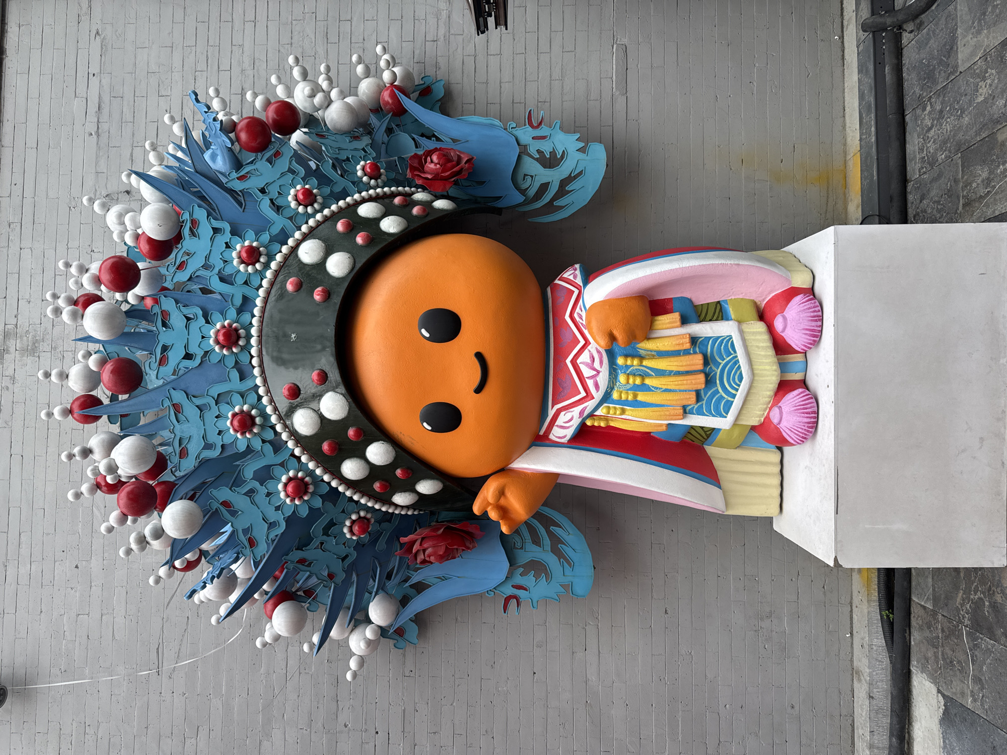 An outdoor image depicts a cartoonish figurine in the style of traditional Chinese opera. The figure has an orange, spherical head with two black, round eyes and a simple, curved smile. It wears a black headdress adorned with rows of white, red, and silver orbs, as well as decorative blue petal-like structures. The figure is dressed in colorful robes with intricate designs and appears to be mounted on a rectangular white stand. The background is a grey brick wall. The subject is in Dongcheng District, China.