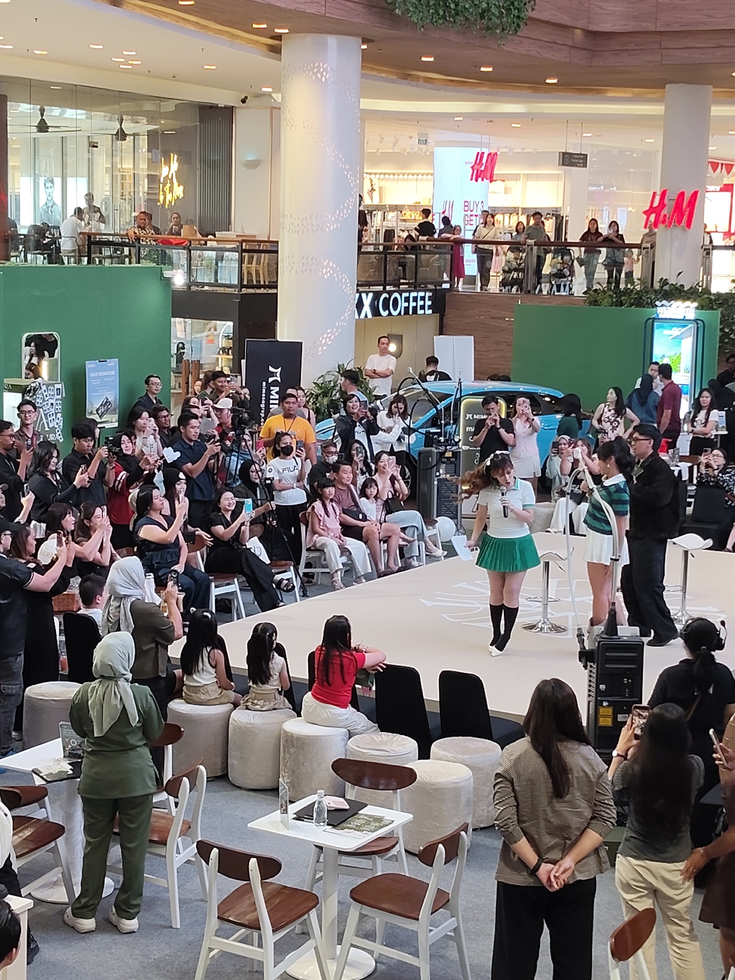The image depicts a lively indoor event, likely a promotional or fashion show, taking place in a shopping mall in Medan, Indonesia. In the foreground, a runway or stage is set up, with several young women dressed in stylish outfits. One woman in a white polo shirt and a green pleated skirt is holding a microphone and speaking, suggesting she is a performer or host. She is flanked by other women in similar attire, one in a green and white striped top and a white skirt, and another in a green top and white skirt.
The stage area is surrounded by an audience seated and standing, many of whom are actively taking photos or videos with their smartphones. This audience comprises a diverse group of people of various ages and genders, dressed in casual to semi-formal attire. The background reveals the characteristic architecture of a modern shopping mall, with tiered levels, large pillars, and storefronts. Signs for brands like H&M are visible, confirming the commercial setting. Natural and artificial lighting illuminates the scene, highlighting the activity and creating a vibrant atmosphere. The presence of a blue car behind the performers suggests it might be part of a product display or stage prop. A few individuals in the audience are wearing hijabs, indicating the cultural context of Indonesia. The overall scene conveys a sense of public gathering and engagement, common in urban shopping centers.