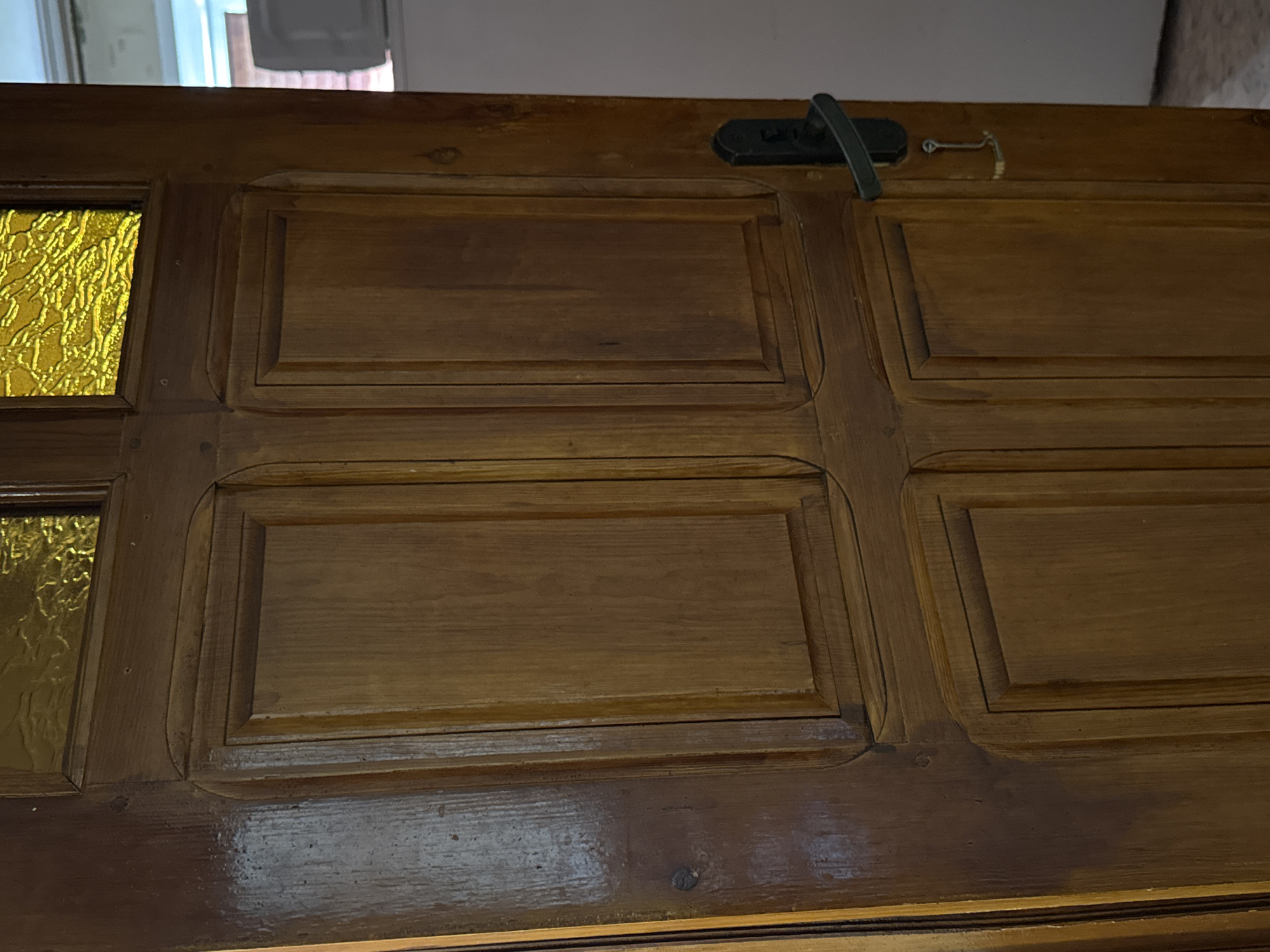 A wooden door with four panels is the main subject of the image. The door is made of dark wood and has a decorative trim around each panel. The top two panels have glass inserts with a textured yellow pattern. A black door handle is visible on the right side of the door, and a small metal latch with a chain hangs from it. The door is partially open, revealing a glimpse of what appears to be a refrigerator or appliance in the background. The lighting suggests an indoor environment. The specific location of Яромінскі сельскі Савет, Belarus is not visually indicated in the image.