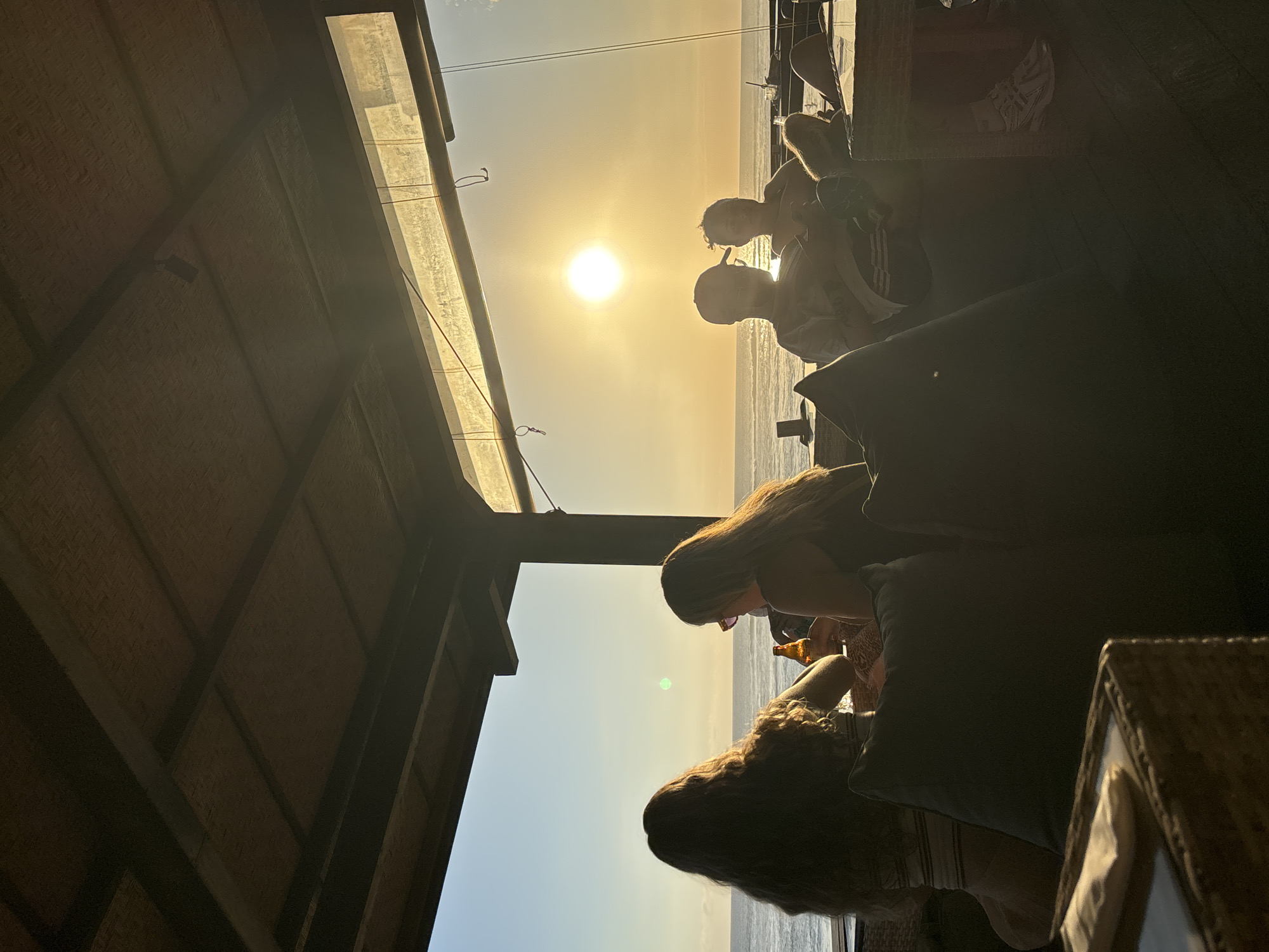 Captured in North Kuta, Indonesia, the photograph depicts a group of individuals gathered at what appears to be an outdoor lounge or bar area, likely positioned near the sea. The setting sun is visible on the horizon, casting a warm glow across the scene. People are seated on cushioned furniture, and some are holding beverages. Overhead, a thatched or textured roof structure provides shade. The image orientation is rotated, presenting an upward view of the scene.