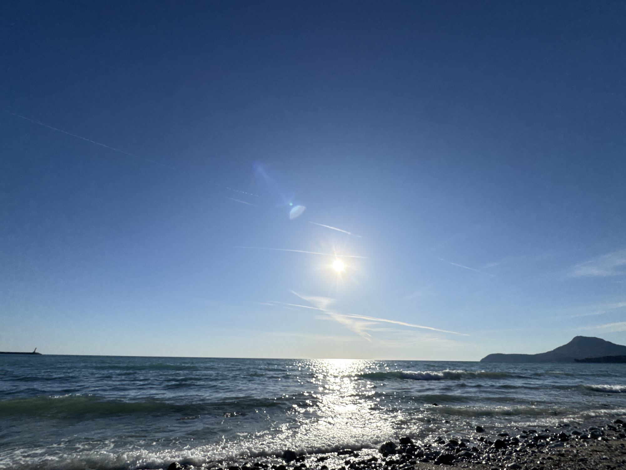 This image presents a serene coastal landscape captured on a bright, clear day in Bar, Montenegro. The scene is dominated by the sparkling sea and a vast, cloudless blue sky.
The sun is prominently featured in the upper-middle section of the sky, emitting a strong, starburst-like light with visible lens flare. Its intense rays create a brilliant, shimmering path of reflections stretching across the water's surface towards the viewer. Several faint, white contrails from aircraft are visible against the deep blue canvas of the sky, suggesting clear atmospheric conditions.
The sea exhibits gentle waves, with white foam visible as they break on the shore in the foreground. The water appears a deep blue-green, reflecting the bright sky. The immediate foreground consists of a pebbly beach, composed of dark, smooth stones glistening from the water.
In the distance on the left, a long, low structure, possibly a breakwater or pier, extends into the sea, topped by a small, dark object that could be a lighthouse. On the right side of the horizon, a distinct, dark-silhouetted mountain or large landmass rises from the sea, adding a dramatic geological element to the composition.
The image conveys a sense of peaceful natural beauty and bright, sunny weather. No people or visible text are present. The time of day appears to be mid-day, given the high position and intensity of the sun.