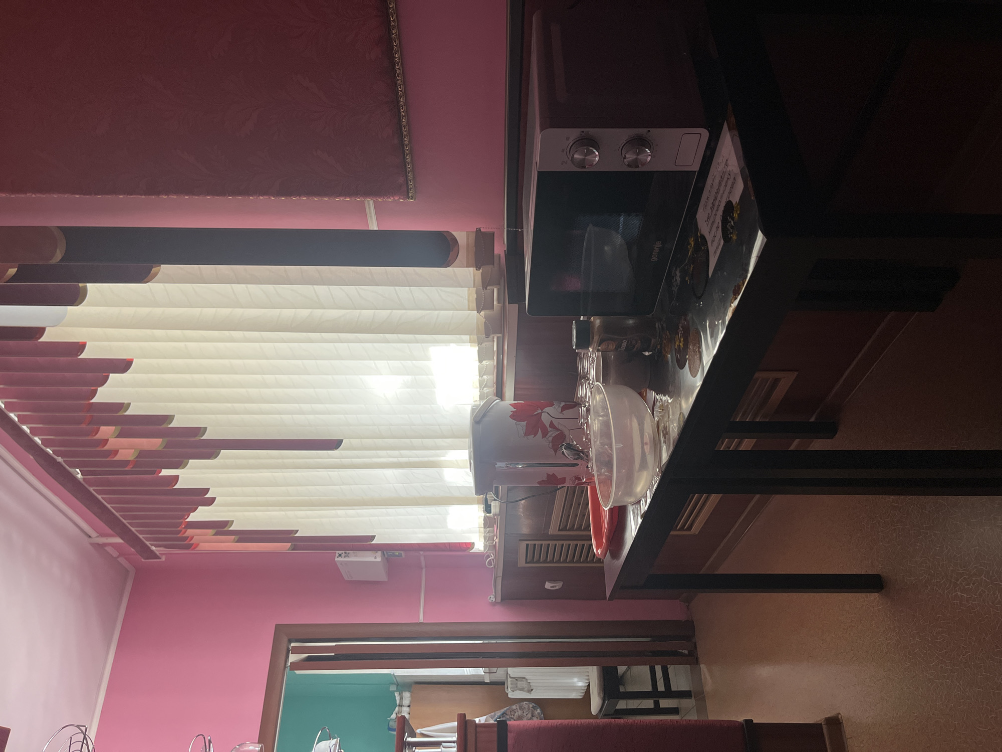 The image depicts an indoor scene, likely a kitchen or dining area, with bright pink walls and a large window covered by vertical blinds. A dark-colored table sits in the foreground, featuring a microwave, a clear bowl, and some jars. In the background, a large appliance with a floral pattern is visible, along with a red tray and glassware. The room also contains a doorway leading to another area, a radiator, and a wall-mounted unit. The lighting suggests it is daytime, with light filtering through the blinds. The overall impression is of a lived-in domestic space. The location is specified as Sorochinsk, Russia.