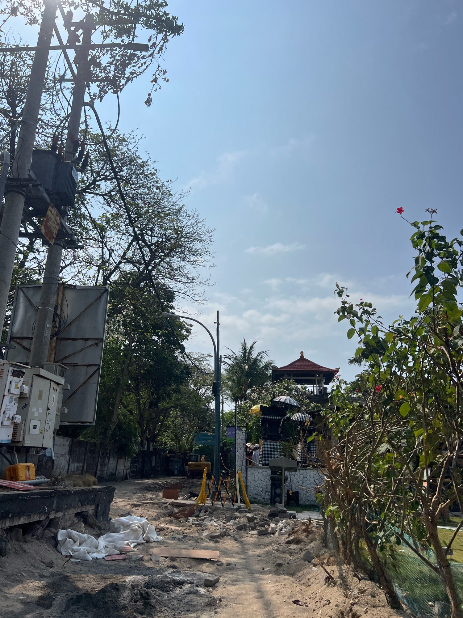 The image shows a street scene in Kuta, Indonesia, with ongoing construction. A power pole with a transformer and electrical equipment is prominently featured on the left. The road is unpaved and appears to be under repair, with piles of sand, debris, and construction materials visible. In the background, a traditional Balinese building with a red roof and intricate details stands out, surrounded by trees and foliage. The sky is clear with some scattered clouds. There are no visible people actively working on the construction site in this frame, but there are indications of recent activity. A yellow excavator is partially visible further down the road, suggesting heavy machinery has been or is being used. The overall scene depicts a typical day in a developing or undergoing renovation area.