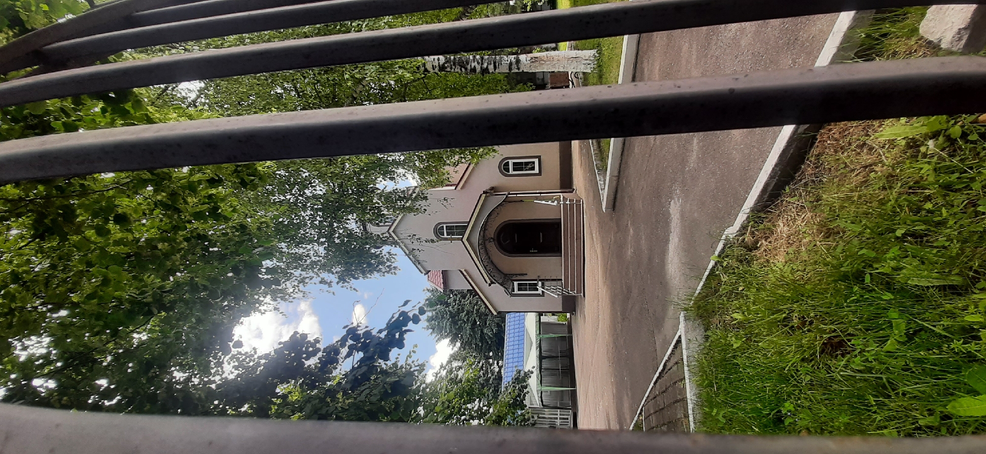 The image captures a small, light-colored church building situated within a fenced-off area. The foreground is dominated by a dark metal fence, partially obscuring the view. Beyond the fence, a paved pathway leads to the church's entrance, which features a covered porch with steps and railings. The church has a simple, symmetrical design with arched windows and a pitched roof. To the left of the church, a structure with a blue, tent-like roof is visible. Lush green trees with dense foliage surround the church, with sunlight filtering through the leaves, creating dappled light on the ground. The sky above is partly cloudy. The scene appears to be a place of worship in a tranquil, possibly rural or suburban setting in Yessentukskaya, Russia.