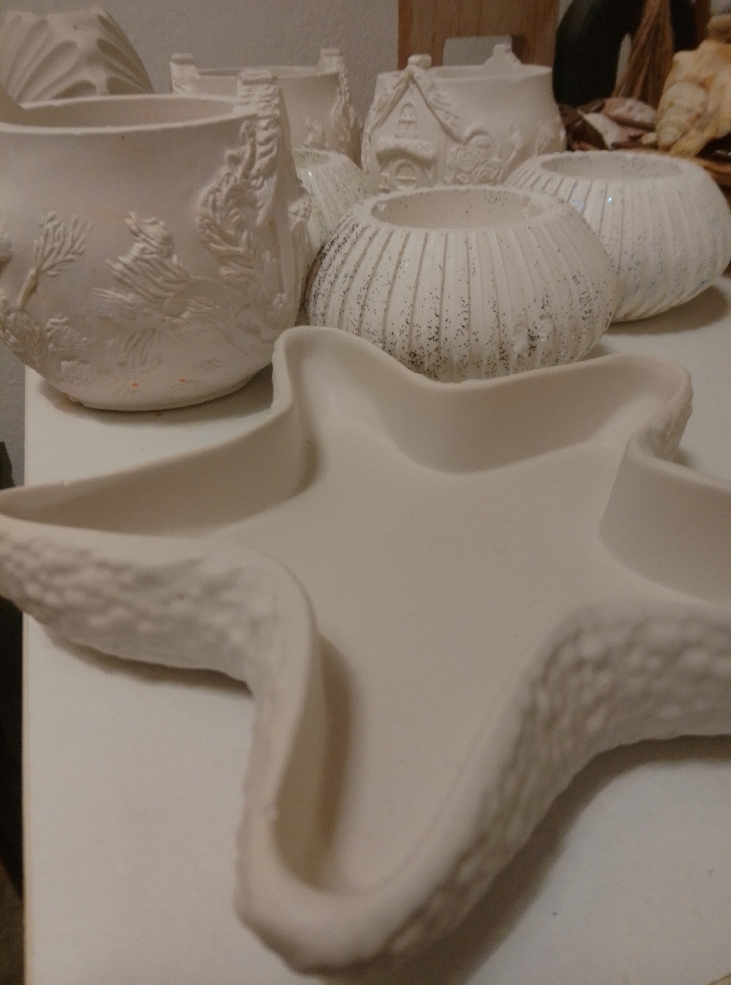 The image shows an assortment of unpainted ceramic items, likely prepared for pottery painting or ceramic glazing. In the foreground, a large, off-white ceramic dish shaped like a starfish dominates the view. Its surface is smooth with textured edges resembling small bumps. Behind it, several other ceramic pieces are visible. To the left, a large planter or vase with intricate embossed detailing of trees and possibly animals is present. To its right, a smaller, round ceramic piece decorated with vertical ridges and flecks of glitter can be seen. Further in the background, another similar ridged ceramic item is partially visible. A ceramic piece resembling a small house with a peaked roof and windows is also in the background. Other unpainted ceramic objects, including a curved shell-like item and a cylindrical pot, are scattered throughout the scene. The items are placed on a plain, white surface. The lighting is soft and diffused. There are no people visible in the image. The scene depicts a collection of unfinished ceramic art pieces, possibly in a studio or home setting. There is no information in the image to determine its location in the city of Wedel, Germany.