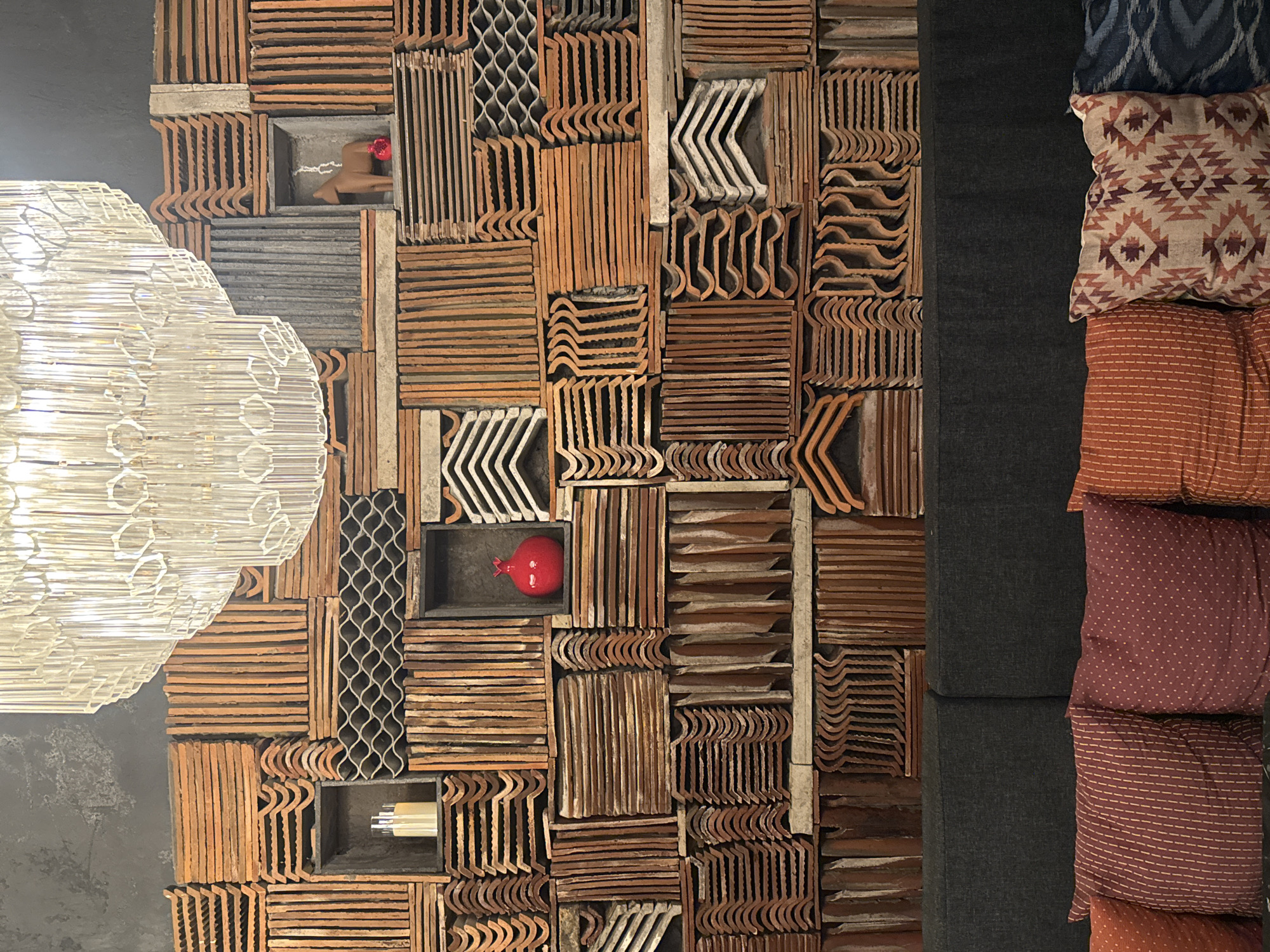 The image shows a close-up of an interior wall featuring a decorative arrangement of terracotta roof tiles of various shapes and sizes. These are interspersed with recessed niches containing objects such as a wooden bird figurine with a red flower, a red pomegranate, and white cylindrical objects. To the left of the wall is a portion of a decorative light fixture with a round shape. To the right is a section of a dark gray upholstered bench or seating area with several decorative pillows in various patterns and colors. The photograph appears to be taken indoors in the Ubud District, Indonesia.