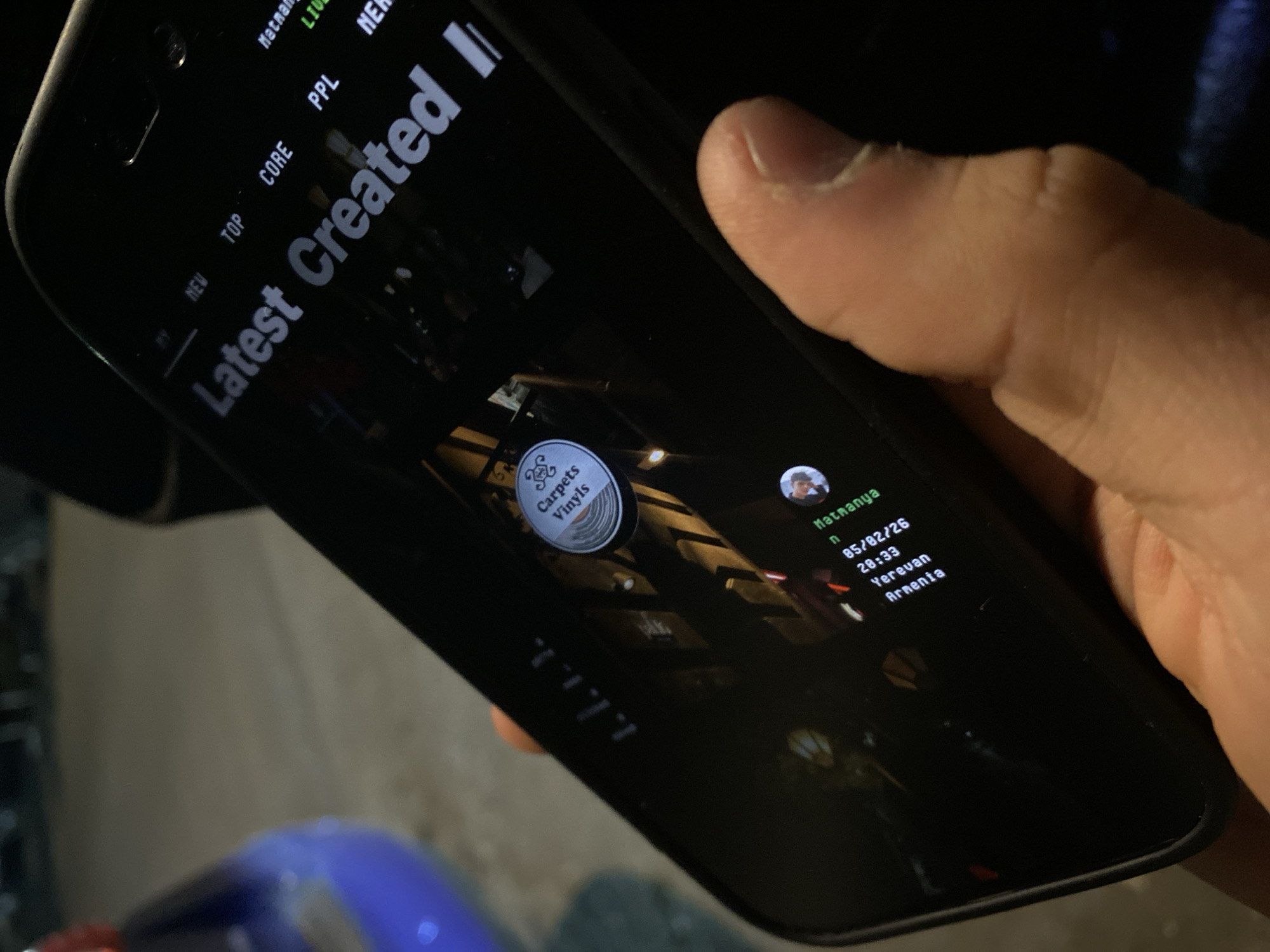 A hand holds a smartphone displaying a social media app. The screen shows a "Latest Created" feed with an album cover and user details: "Matmanya n 05/02/26 20:33 Yerevan Armenia". The phone is held at an angle, with the city environment barely visible in the background.