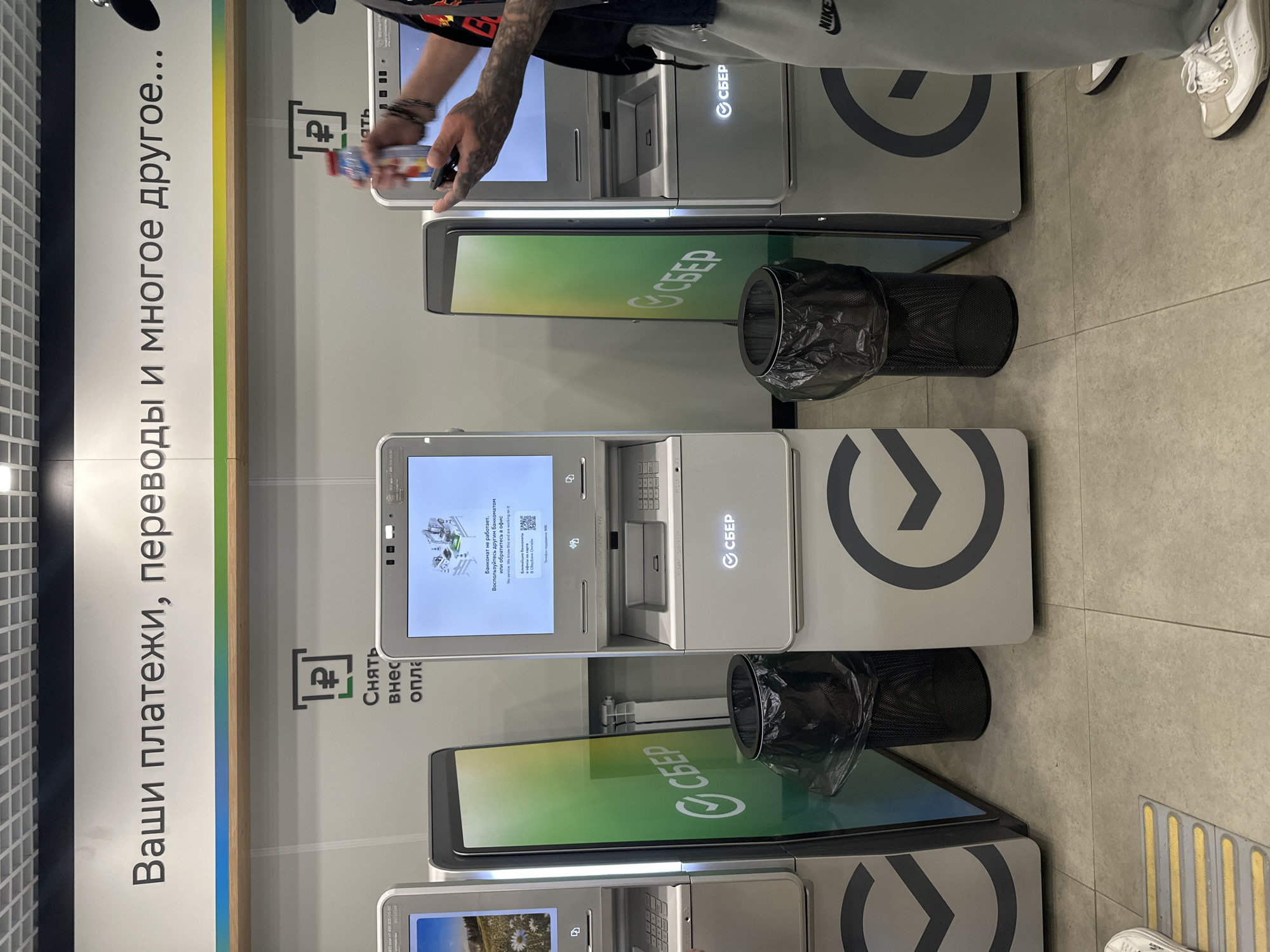 The image shows a row of Sberbank ATMs in what appears to be a public indoor space. On the far left, a partial view of an ATM with a screen displaying a natural landscape is visible. Next to it is a green Sberbank branded standee with a trash can in front of it. In the center, a smaller, white Sberbank ATM is operating, its screen displaying a diagram of an ATM and text in Russian indicating it is out of service and suggesting alternative services. To the right of this is another Sberbank ATM. A person, partially visible, is standing in front of the rightmost ATM, holding a bottle of hand sanitizer and a mobile phone. The floor is tiled, and there is a yellow and grey tactile paving strip in the foreground. The overall environment appears to be a bank branch or a commercial space equipped with ATMs. The sign above the ATMs reads "Ваши платежи, переводы и многое другое..." which translates to "Your payments, transfers, and much more...".
