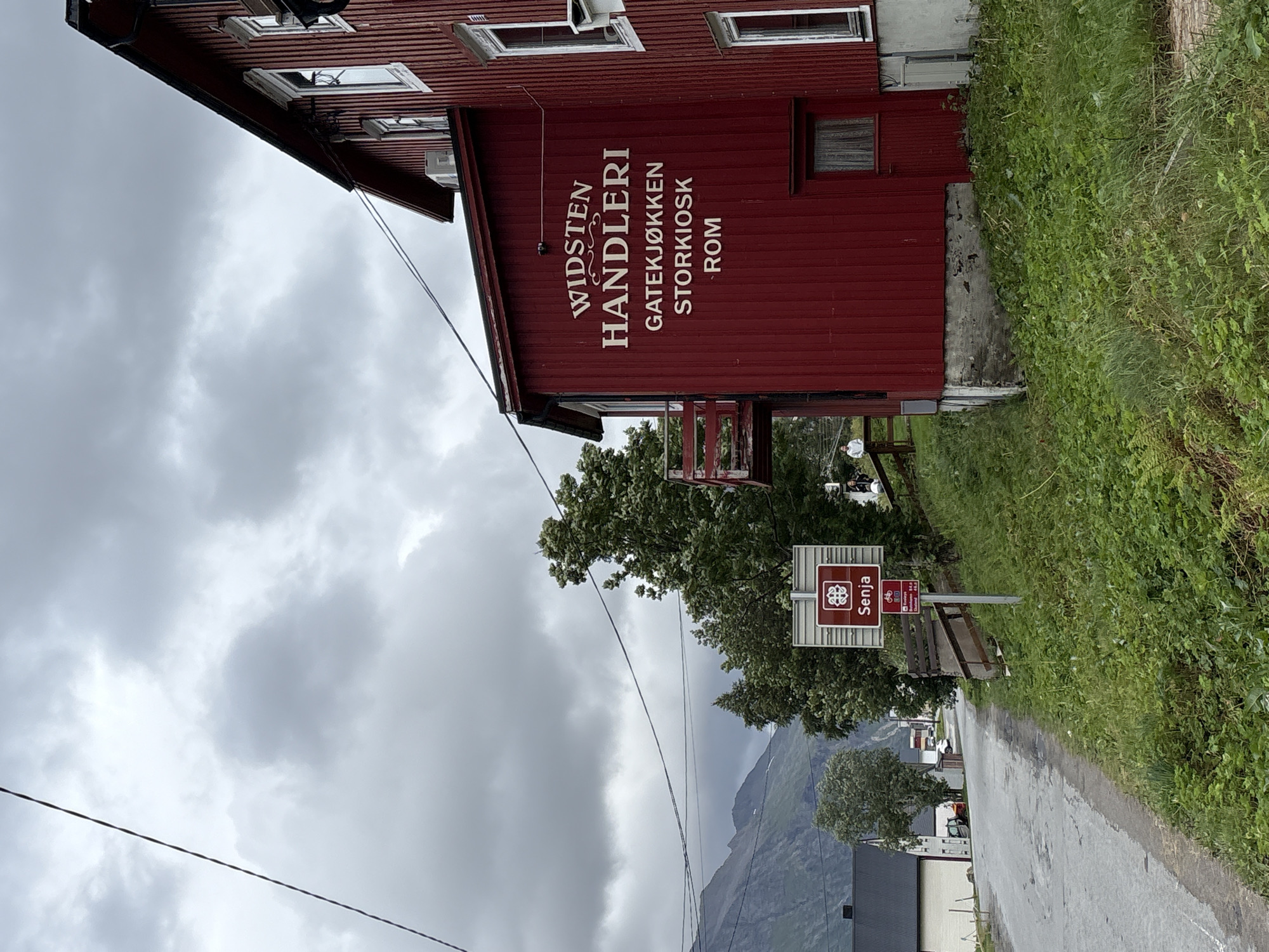 The image captures a cloudy day in Gryllefjorden, Norway, with a prominent red wooden building that houses "WIDSTEN HANDLER", "GATEKJØKKEN", "STORKIOSK", and "ROM". A sign with the word "Senja" is visible to the left of the building, accompanied by cycling and walking symbols. The building is situated on a grassy slope, with a paved road leading away from it and towards distant mountains. Several parked cars and a few people are visible on the road in the background. The overall scene depicts a rural setting with a commercial establishment catering to travelers.