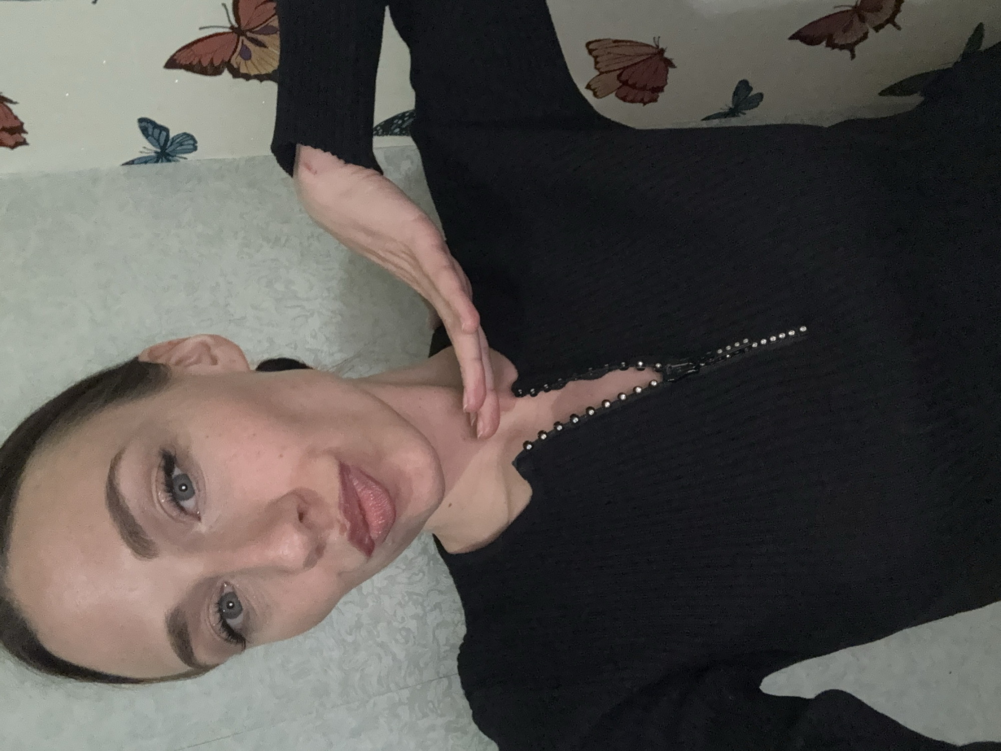 A medium, eye-level shot depicts a woman indoors. Her face is turned toward the camera, resting on a light-colored surface, possibly a bed or floor. She has fair skin, blue eyes, defined eyebrows, and pink lips, and her hair is pulled back. She is wearing a black long-sleeved dress with embellishments along the neckline. Her right hand is raised, fingers extended, near her neck.
The background features a wall covered in wallpaper with a repeating butterfly pattern in various shades of brown and blue. The lighting is soft, casting subtle shadows and highlights across the scene. There is no evidence to confirm the location of Saint Petersburg, Russia.