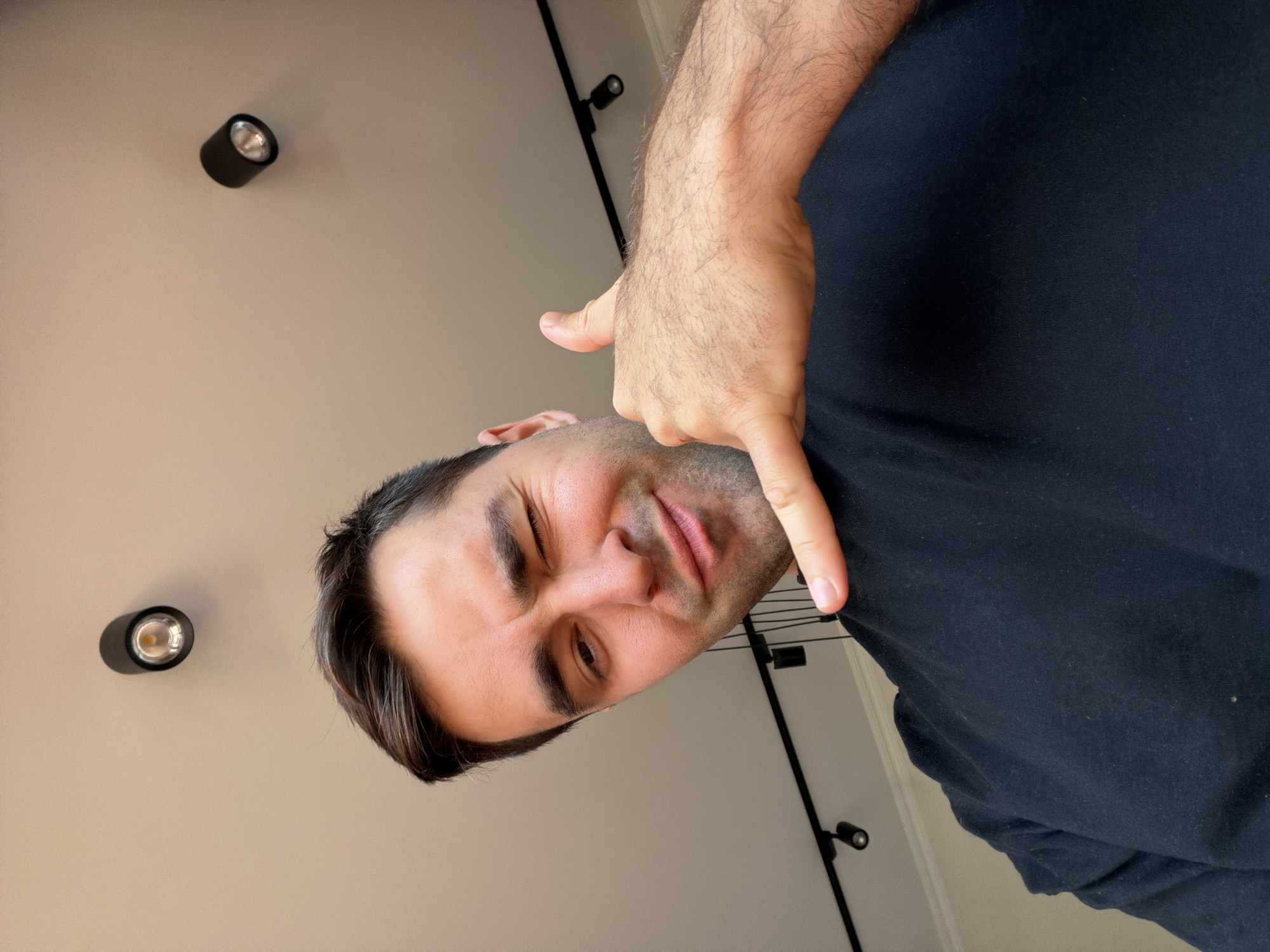 A selfie of a light-skinned man with dark hair is oriented at an angle. The man is winking with his left eye and pointing downward with his right hand. He is wearing a dark-colored shirt. In the background, a light-colored ceiling is visible, with a track lighting system installed. This scene is likely located inside a building in Tbilisi, Georgia, but that cannot be conclusively determined from the image.