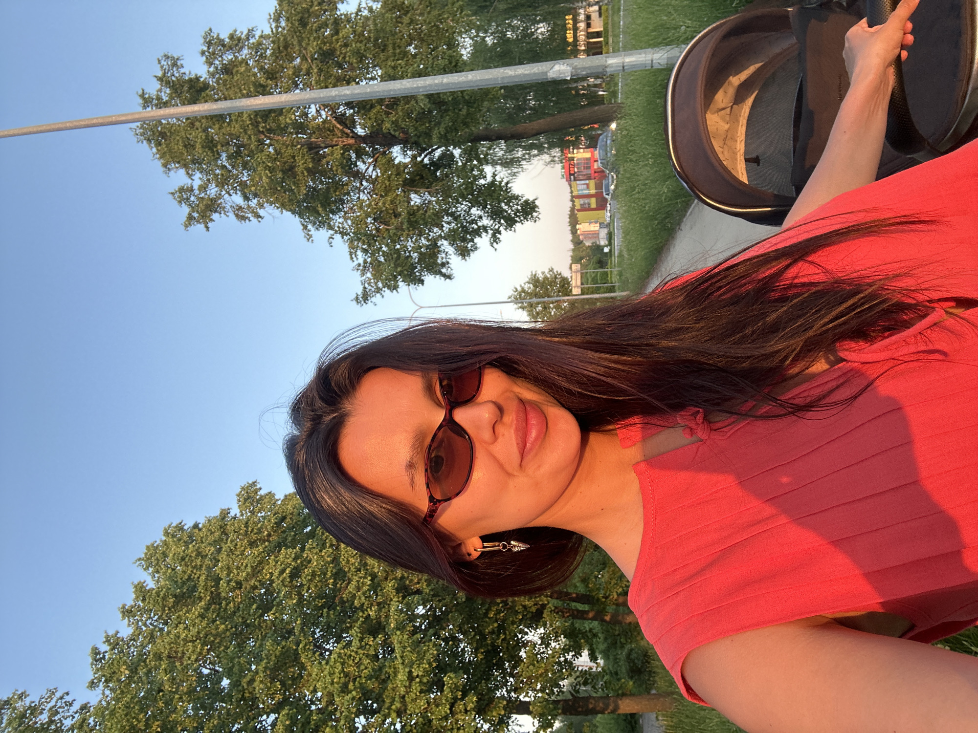 A woman is standing outside next to a stroller. She is wearing sunglasses and a red top. Trees and a blue sky are visible in the background. Some buildings are visible in the distance. The location is Chaikovsky, Russia.