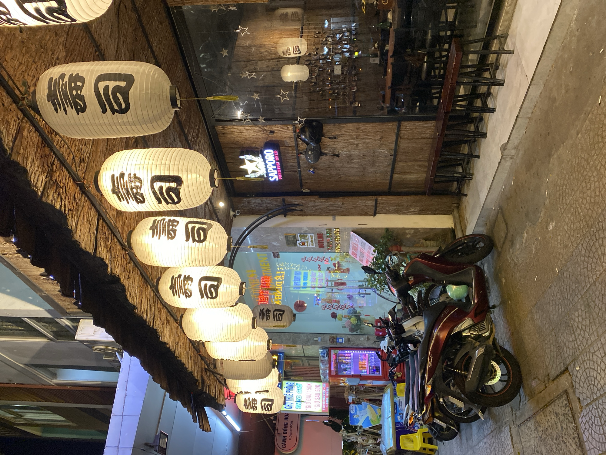 The image captures a street view at night in Đà Nẵng, Vietnam, likely in front of a Japanese restaurant. Several illuminated paper lanterns, adorned with Japanese characters, hang from a thatched roof extension. Below the lanterns, a glass storefront reveals the interior of a restaurant with wooden walls and seating. A prominent "Sapporo Premium Beer" sign is visible on the exterior. To the left, motorcycles are parked on the tiled sidewalk, and a red beverage cooler stands near a shop entrance. A street sign for "Cánh Đồng Hoa" (Flower Store) is also present. The scene is illuminated by the restaurant's lights and the ambient city glow.