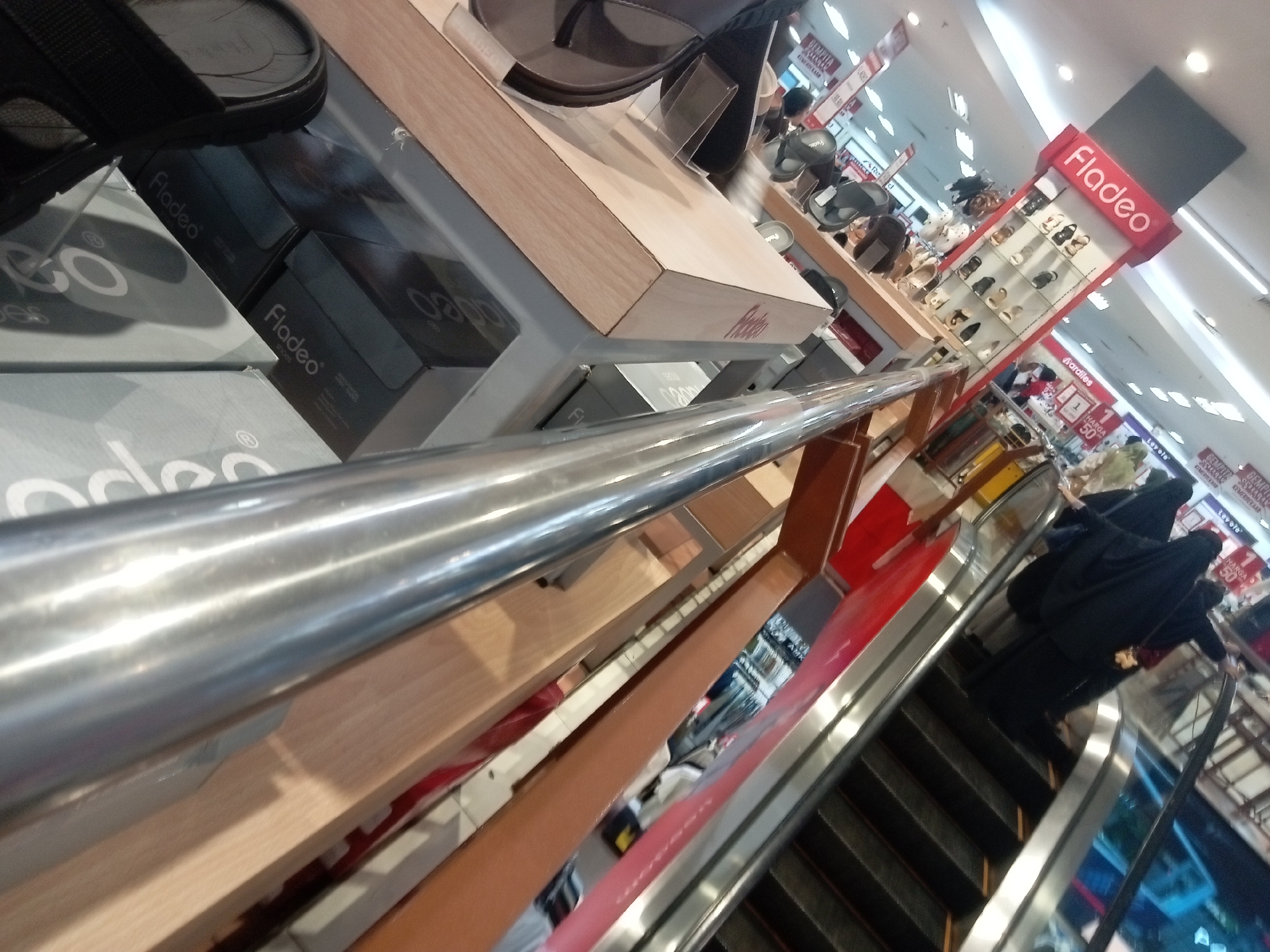 The image depicts the interior of a Fladeo shoe store, likely located in Batam, Indonesia, as indicated by the prominent signage and typical retail environment. In the foreground, a metal railing runs diagonally across the frame, obscuring parts of the store's display shelves and merchandise. Below the railing, several shoeboxes, some labeled "Fladeo," are stacked on a display shelf. The boxes are dark in color, suggesting they contain footwear.
Further into the store, a tiered display unit prominently features the "Fladeo" logo in red. This unit showcases various styles of women's shoes, arranged in neat rows on shelves. The shoes appear to be sandals and flats, in a range of neutral colors.
To the right of the shoe display, an escalator is visible, with people, primarily women dressed in dark, modest attire, ascending it. The escalator's steps are dark, and the surrounding area is lined with polished metal. Behind the escalator, more retail displays and signage are visible, including what appears to be price tags and advertisements, some with red accents. The overall lighting suggests an indoor retail environment, possibly a mall or department store. The image is taken from a slightly elevated, angled perspective, giving a sense of depth and the scale of the store.