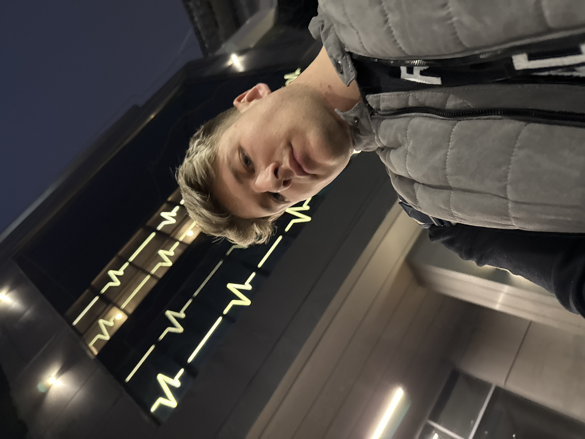 A young male is facing the camera, wearing a gray puffer vest over a black shirt. He has light blonde hair. Behind him, a building facade displays an illuminated zigzag pattern reminiscent of a cardiogram. Based on the visible architectural details and the cardiogram motif, the location is the Medical University of Plovdiv in Bulgaria. The time of day appears to be dusk or evening.
