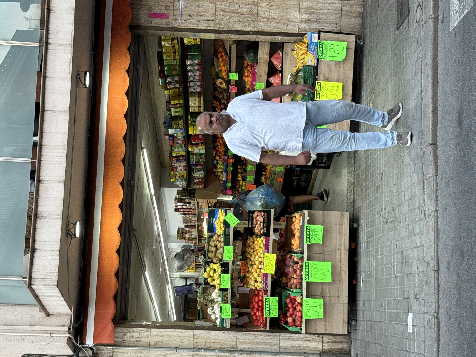 An outdoor market stall in Blanes, Spain, displays a variety of fresh produce, including tomatoes, apples, peaches, nectarines, apricots, melons, bananas, and other fruits and vegetables. The stall is set up in front of a shop with an orange awning. A man in a white polo shirt and light blue jeans is walking away from the camera, towards the right side of the frame, with his hands in his pockets. He is a middle-aged man with dark hair and a dark complexion. Behind him, another person, possibly a shop employee, is visible inside the stall, carrying a blue plastic bag. Several green and yellow signs with prices are displayed on the stall, indicating offers for different fruits. The ground is paved with gray square tiles, and a street curb is visible in the foreground. The overall scene depicts a typical street-level retail environment in a Spanish town.