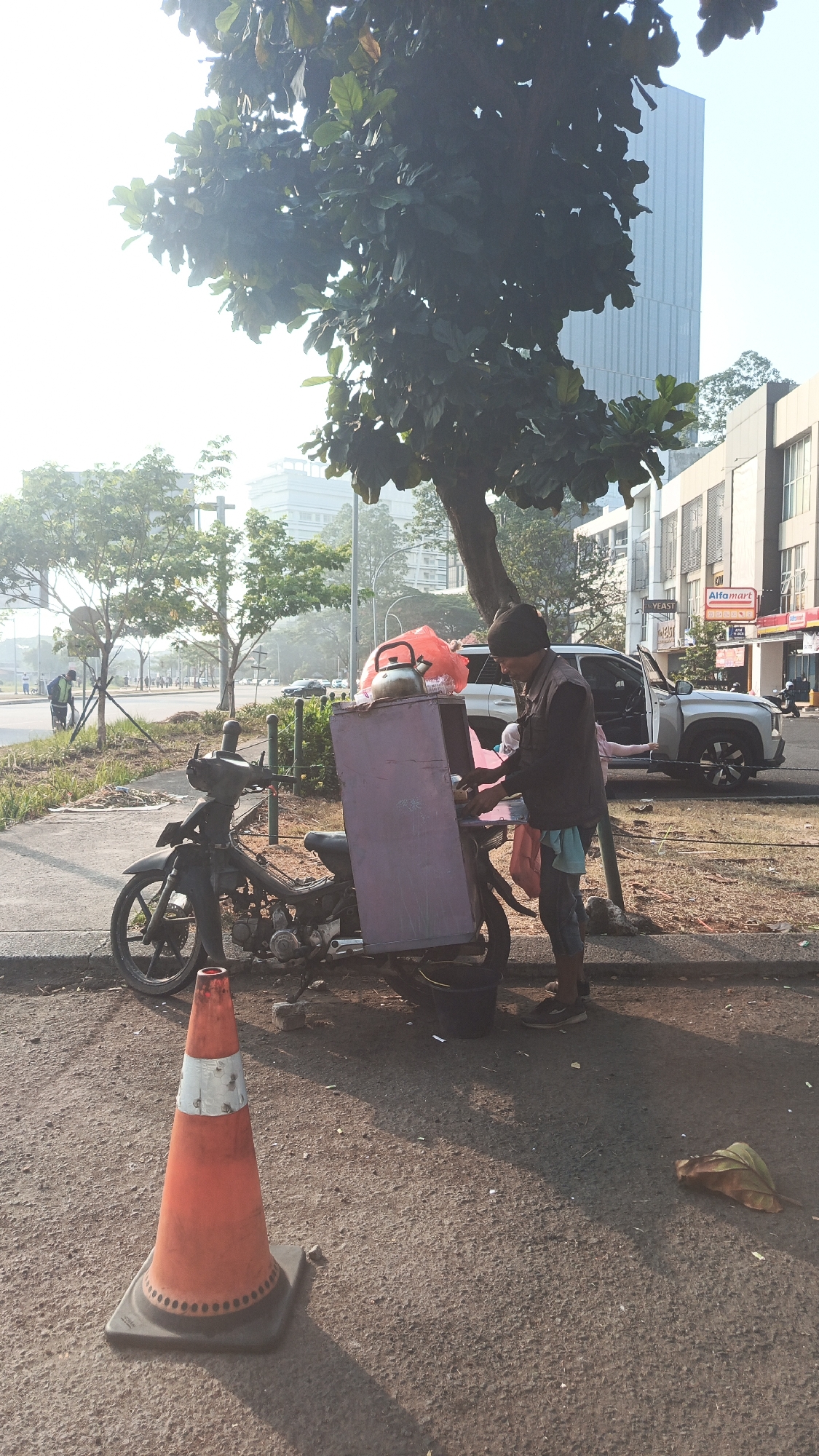A man stands beside a motorcycle with a large purple box attached to the back, in Tangerang, Indonesia. The motorcycle is parked on the side of a road, near a sidewalk. The man is wearing a dark jacket and light-colored pants, and appears to be interacting with the contents of the box. A red traffic cone is in the foreground. In the background, there are trees, buildings, and vehicles on the road. The sky is bright and hazy, suggesting it might be morning or late afternoon. The general environment appears to be an urban or suburban setting with a mix of residential and commercial structures.