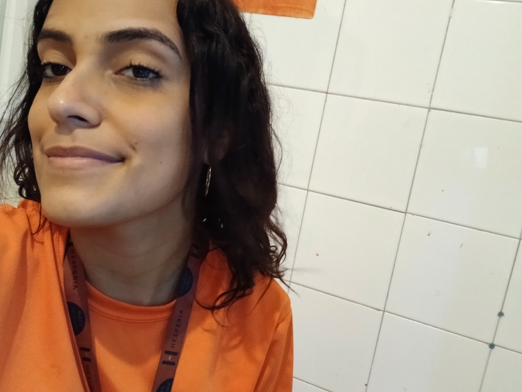 A close-up portrait features a young woman with dark, curly hair and dark eyes, looking slightly to her left with a subtle, gentle smile. She appears to be in her late teens or early twenties, with well-defined eyebrows. A small, golden hoop earring is visible on her left ear. She is dressed in a bright orange long-sleeved top, possibly athletic wear, and wears a dark lanyard around her neck. The lanyard clearly displays the word "HESPERIA" in white text, repeated along its length, accompanied by a stylized 'H' logo.
The background is a white tiled wall with dark grout lines, typical of an indoor or semi-enclosed utility space such as a bathroom or service area. A partial view of another orange object or fabric is visible in the upper right background. The lighting is soft and even, suggesting an indoor setting. The woman's expression conveys a calm and contented mood. Given the location context of Playa El Agua, Venezuela, the setting with the tiled wall and the "HESPERIA" lanyard likely indicate an association with the Hesperia hotel chain, which has resorts in coastal regions, including those near Playa El Agua. The image captures a simple portrait or selfie of the individual.