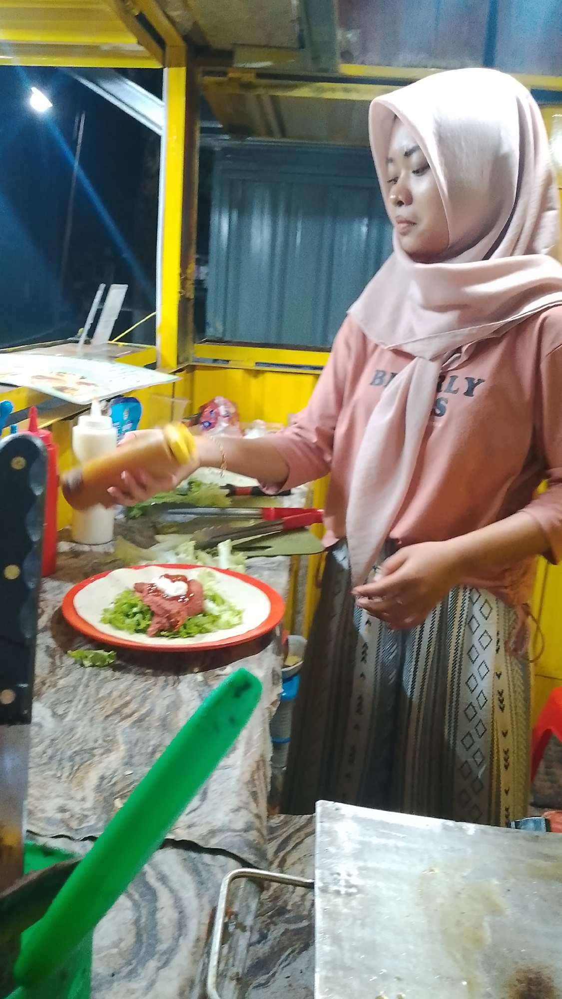 In the city of Karanglangu, Indonesia, a young woman wearing a light pink hijab and a pink t-shirt is standing behind a food stall. She is preparing food, holding a bottle of sauce in her right hand and about to drizzle it onto a wrap on a red and white plate. The wrap contains lettuce, a piece of meat, and a dollop of white sauce. In the background, there are yellow metal structures of the food stall, with a grey corrugated metal wall behind them. Various condiments, including red and white squeeze bottles, are visible on the counter. A green utensil is also in the foreground. The scene appears to be at night, illuminated by artificial lighting.