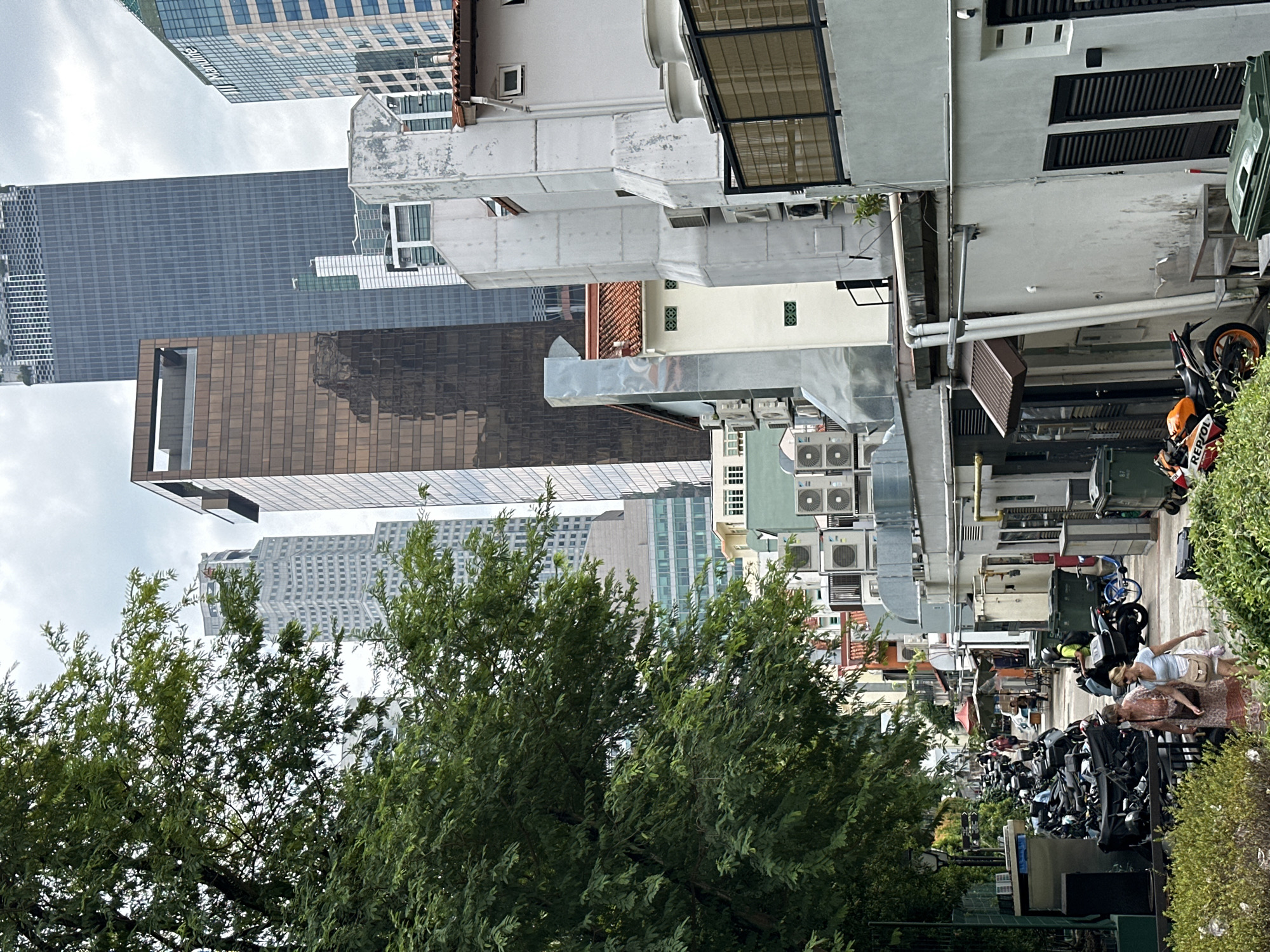This image depicts a street scene in Singapore, likely within the bustling central business district or a nearby heritage area, given the juxtaposition of modern skyscrapers and older shophouse architecture. On the left side of the frame, a lush green tree dominates, its branches partially obscuring the view of the background. Behind the tree, a tall, light-colored skyscraper with a patterned facade is visible. Further back, a prominent dark grey skyscraper rises, its sheer surface reflecting the cloudy sky. To the right of this, a copper-colored skyscraper with horizontal bands is also present.
The right side of the image features a row of lower-rise buildings, characteristic of shophouses, with several air conditioning units mounted on their facades. These buildings are painted in various muted colors, including white and a pale green. The architecture suggests a mix of functional and residential spaces. In the foreground, two women are walking away from the camera, dressed in casual summer attire. They are walking on a paved pathway that runs alongside the shophouses. Beyond them, a street is lined with numerous parked motorcycles, indicating a common mode of transportation in the area. Further down the street, several open-air stalls with umbrellas are set up, suggesting a market or food vendors. A person wearing a yellow vest is seen riding a motorcycle in the middle ground. The sky is overcast with a moderate amount of clouds. The overall impression is one of a vibrant urban environment where old and new coexist.
