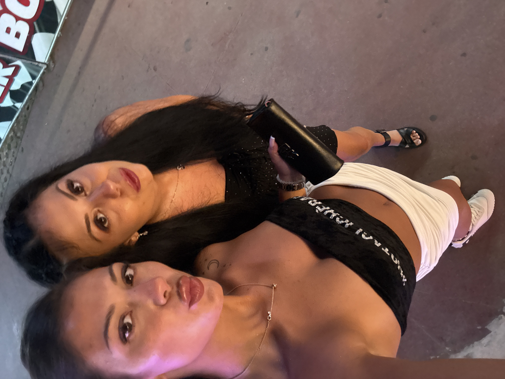 Two women are posing for a selfie outdoors on a paved street. The woman on the left is closer to the camera, with her lips puckered. She is wearing a black bandeau top and a white skirt. A thin, gold necklace with small charms is visible on her neck. The woman on the right is slightly behind and to the right of the first woman. She has long, dark hair and is wearing a black dress. She is holding a black clutch bag in her right hand. In the background, a sign with red and white lettering is partially visible. The lighting suggests it is evening or nighttime. Based on the architectural style suggested by the sign in the background (which appears to be part of a fairground attraction typical of tourist areas), and the presence of an inscription in Cyrillic script on the bandeau top, the image was likely taken in a tourist area of Nesebar, Bulgaria.