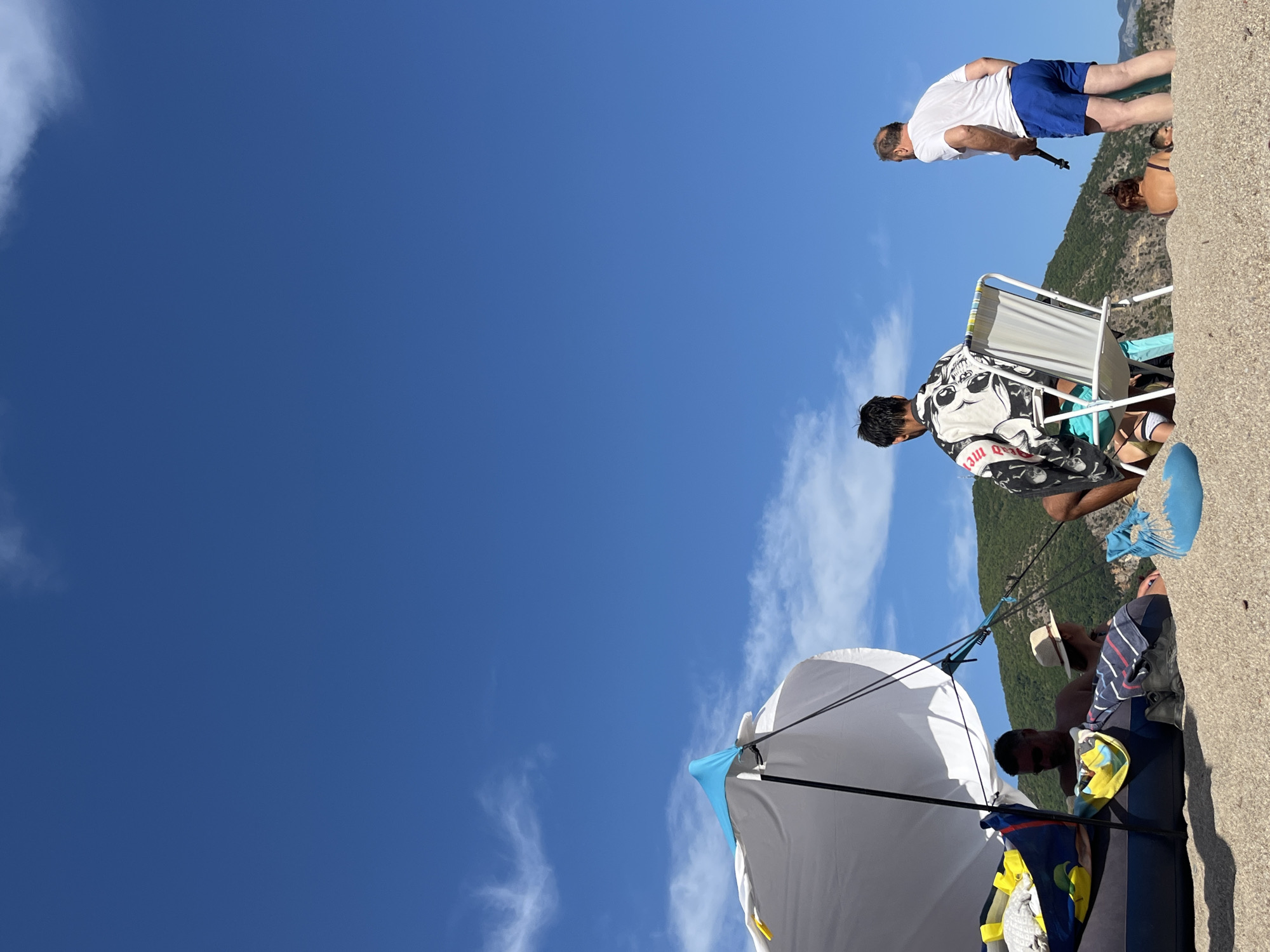 The image captures a serene beach scene under a bright blue sky with sparse white clouds. In the foreground, on a sandy beach, a group of people are relaxing. A large, white beach tent with blue accents provides shade for some. A man wearing sunglasses and a striped towel is seated on an inflatable structure within the tent. Next to him, a person wearing a straw hat is also seated. To the right of the tent, a young person is seated in a white lawn chair, wearing a distinctive black and white patterned shirt and turquoise shorts. Further to the right, an adult male in a white t-shirt and blue shorts stands holding a dark object, possibly a camera or a stick. In the background, a person is partially visible sitting in the sand. Beyond the beach, lush green mountains rise against the clear sky, suggesting a coastal or mountainous region. The overall atmosphere is one of leisure and outdoor recreation. The location is the city of Prijevor, Montenegro.