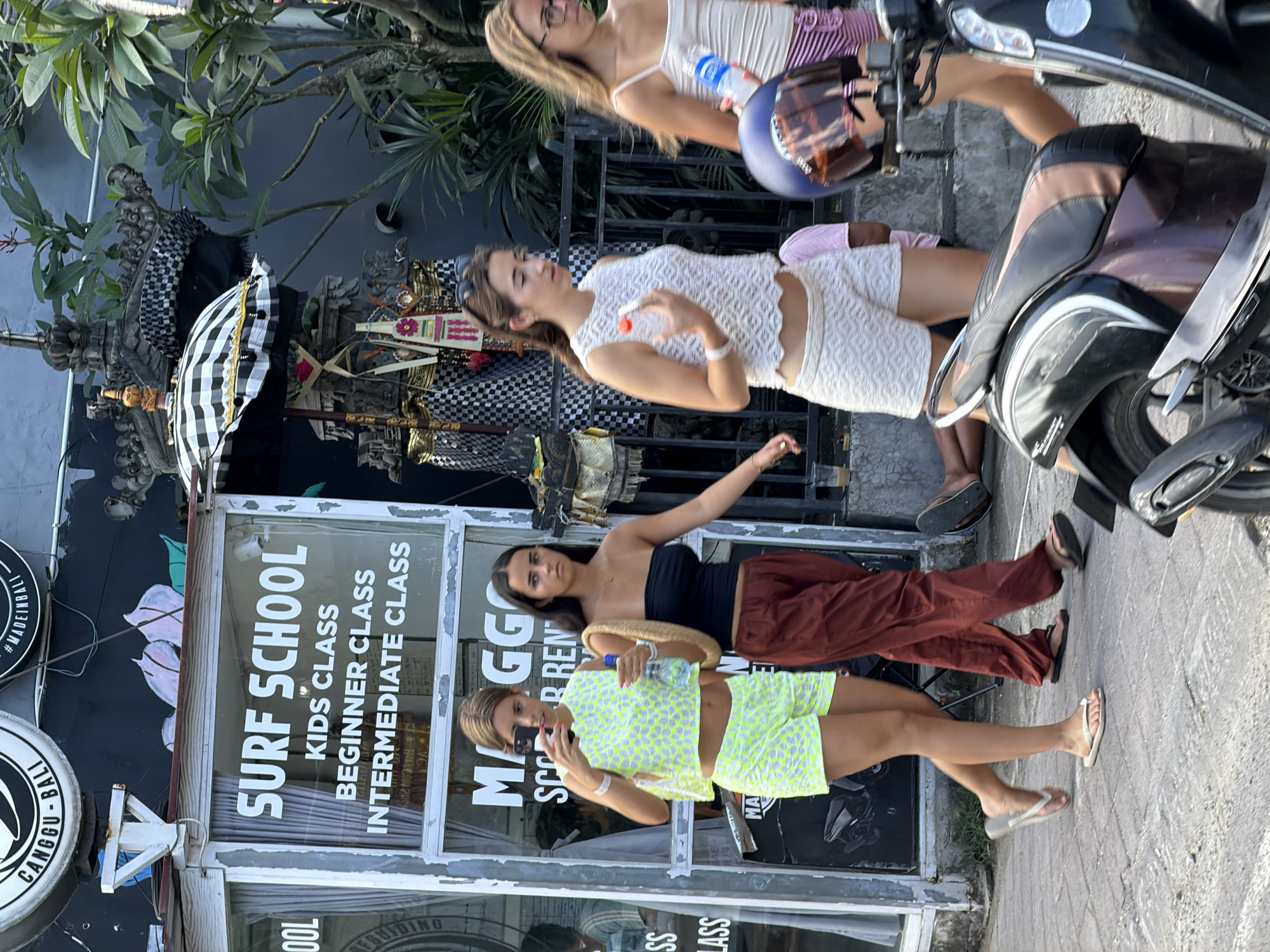 In North Kuta, Indonesia, three young women walk past a surf school. The woman on the left wears a neon yellow patterned crop top and shorts, holding a phone up as if taking a selfie. The woman in the middle is dressed in a black tube top and rust-colored pants, carrying a straw bag over her shoulder. The woman on the right wears a white lace top and shorts, holding a bottle of water. A motorbike is parked in the foreground on the right, with a helmet on the handlebars. The background shows a building with "SURF SCHOOL" visible on the window, along with smaller text listing classes. Traditional Balinese decorations are also visible behind the women. The pavement is made of grey cobblestones, and foliage can be seen behind the building. The scene appears to be a street during daylight hours.
