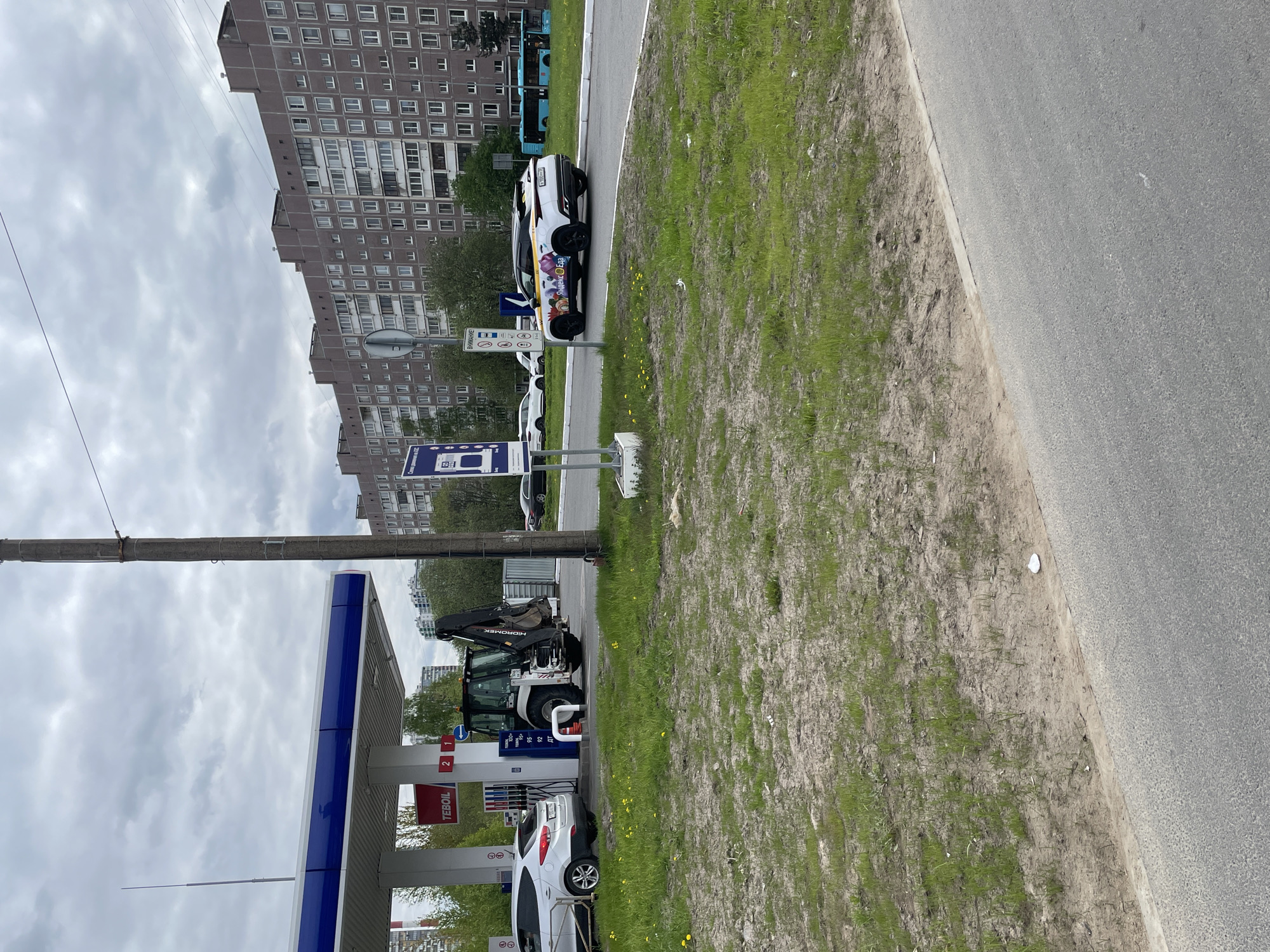 The image depicts a street scene in Saint Petersburg, Russia. A TEBOIL gas station is visible on the left side, along with a white car and a piece of heavy construction equipment parked in front. Next to the road is a grassy area, followed by the asphalt road. Further along the road, a white car with advertisements parked. In the background, there is a large apartment building with rows of windows under an overcast sky. The light of the day seems cloudy.