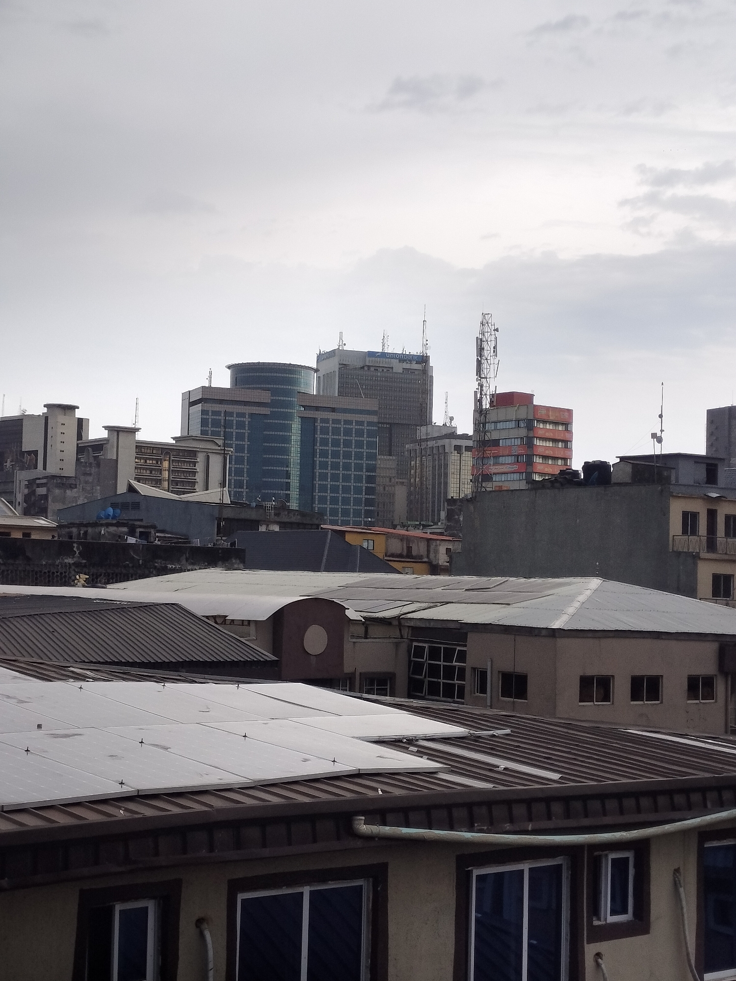 The image depicts a cityscape in Lagos, Nigeria, characterized by a mix of low-rise residential buildings in the foreground and modern high-rise commercial buildings in the background. The sky is overcast, creating a muted lighting effect.
In the foreground, several buildings feature a variety of roof types, including corrugated metal and flat surfaces. Some roofs are equipped with solar panels. The buildings have windows of different sizes and styles, and the exterior walls appear to be painted in neutral tones.
In the background, high-rise buildings dominate the skyline. These structures are predominantly glass and steel, with some featuring distinctive architectural elements, such as rounded facades and antennas. One building is identified with the "Union Bank" logo. A communication tower is visible among the buildings.
The overall impression is that of a densely populated urban area, with a mix of older and newer construction. The location is likely the central business district of Lagos, Nigeria.