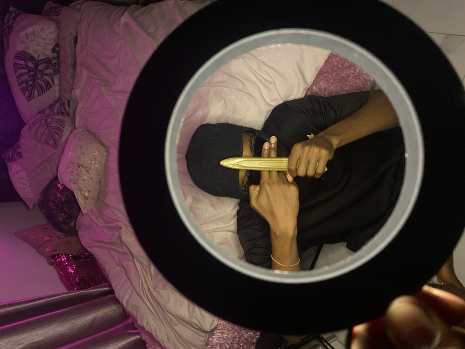 A person wearing a black balaclava and black long-sleeved shirt lies on a bed, viewed through a selfie light. They hold a gold object that resembles a knife or letter opener, resting it on the fingers of their other hand. The bed is covered with a white comforter and multiple pillows, some with printed designs and others with glittery sequins. A pink hue casts over the scene.