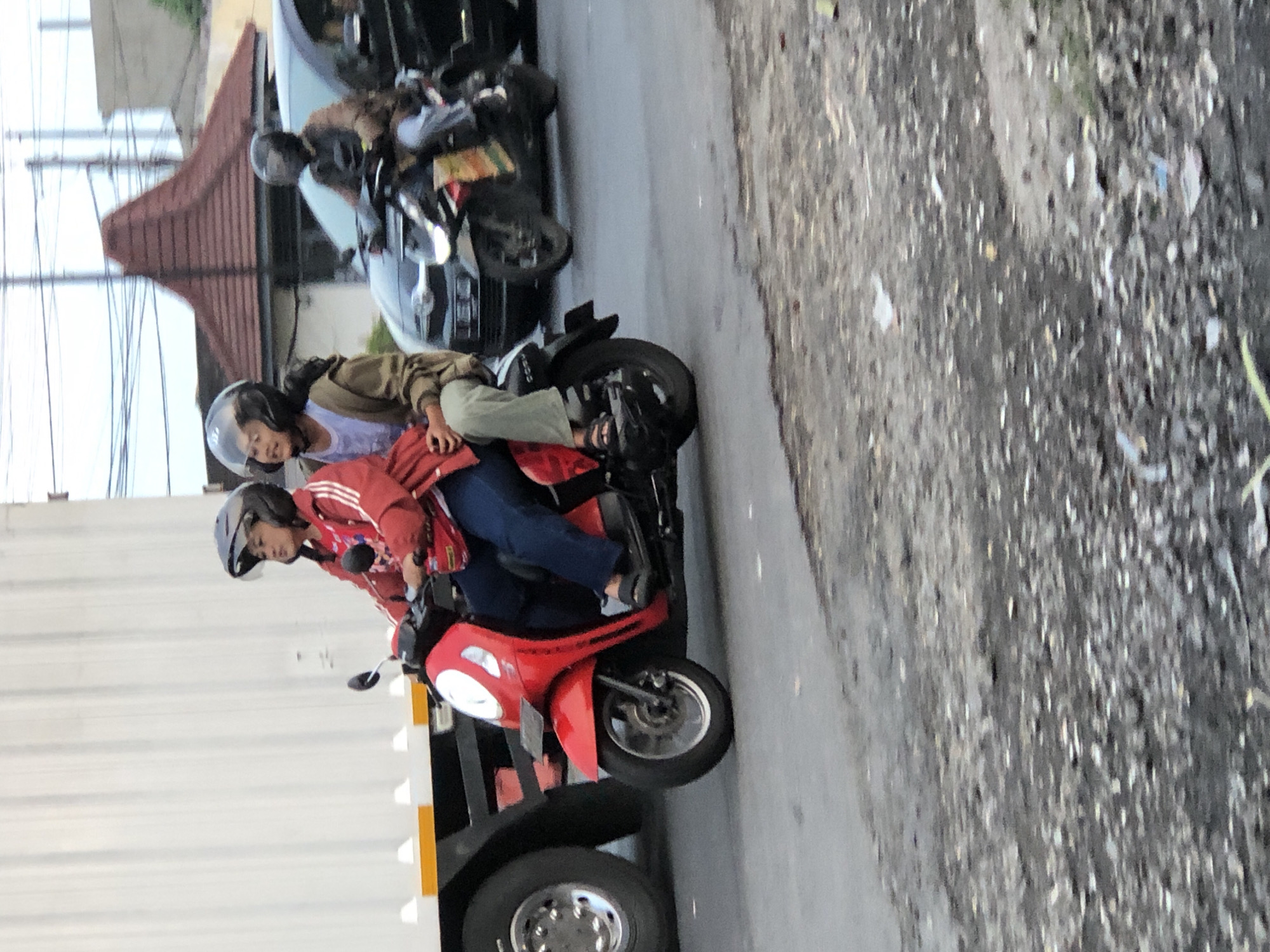 Two individuals, a driver and a passenger, are riding on a red scooter. The driver, wearing a red jacket and a helmet, is looking to the right. The passenger, also wearing a helmet, is looking forward. They are on a road with other vehicles present, including a truck to the left and a black car behind them. The scene appears to be in an urban or semi-urban environment, possibly in Kludan, Indonesia, judging by the context. The road surface is asphalt, with some debris scattered on it. The overall lighting suggests it is daytime.