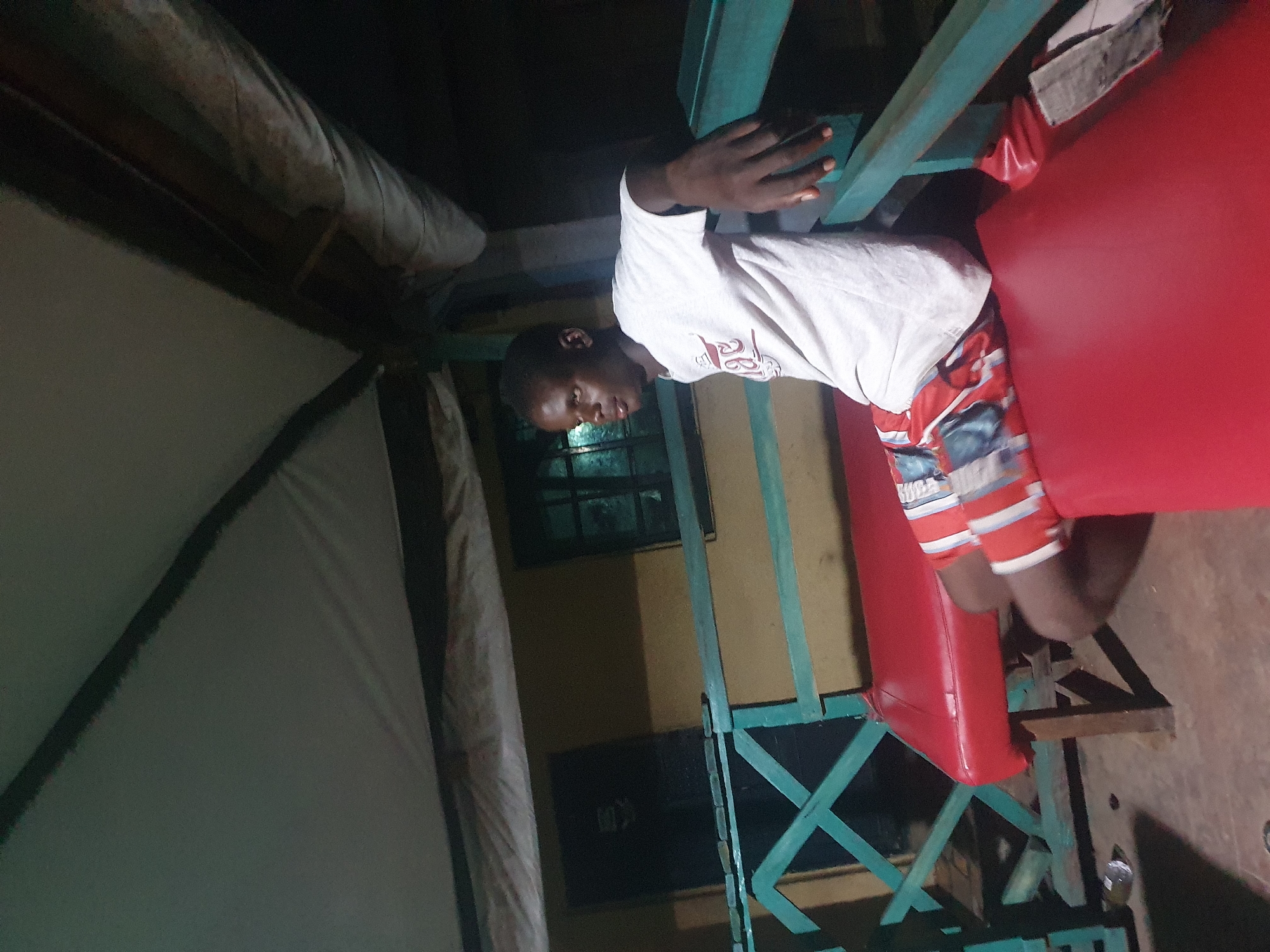 A young Black man wearing a white t-shirt and red and white striped shorts sits on a red bench outside a building. He is leaning on a teal-colored railing and looking to his left. The building behind him has a yellow wall and a window with a dark pane. A dark door is visible to the left of the window. A white awning or canopy is stretched above the scene. The ground is a reddish-brown color.