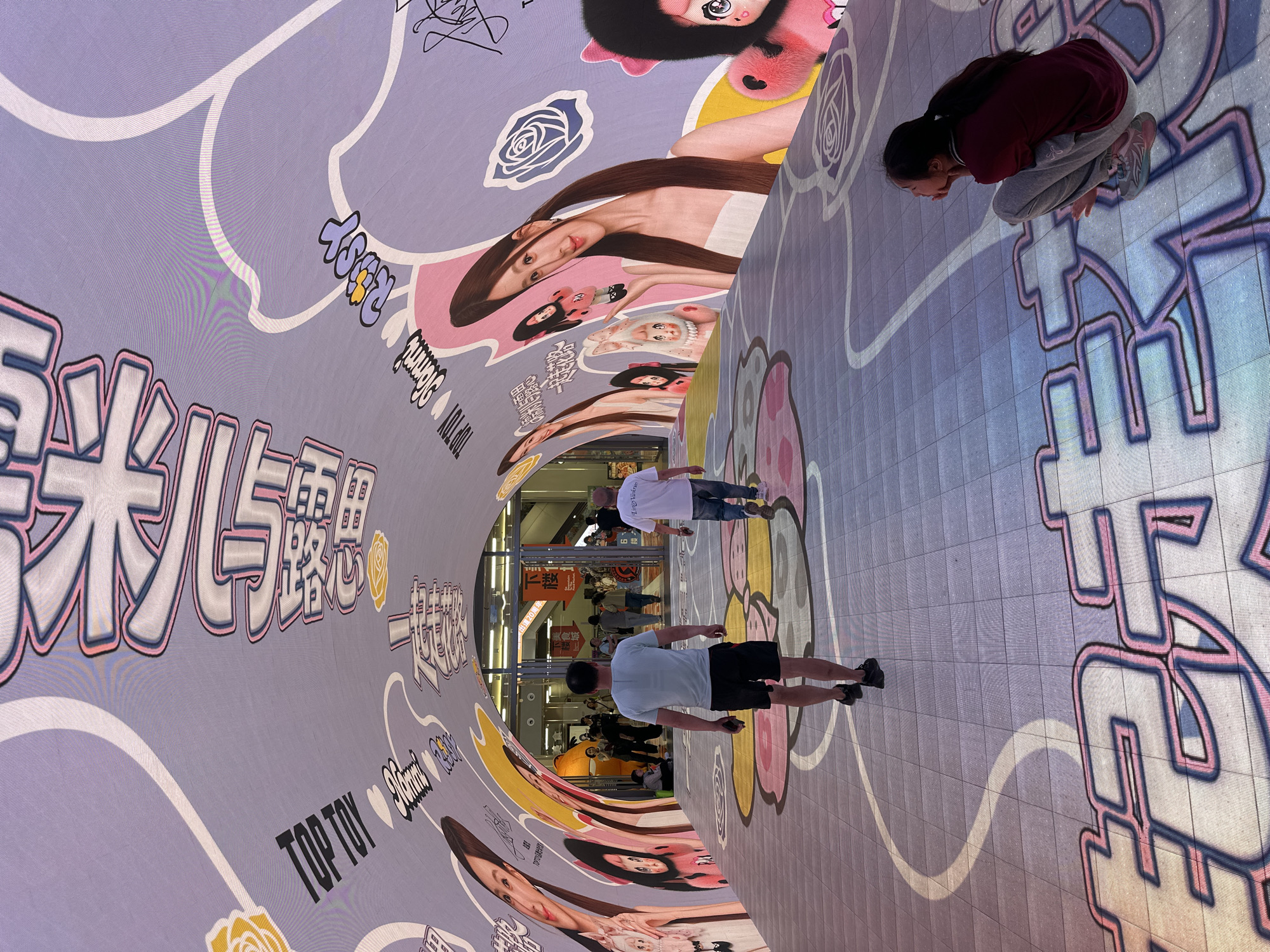 This photograph shows a semi-cylindrical structure with interior advertising, with two people standing inside it and a third crouching. The tunnel-like structure is decorated with images of a woman and branding, and Chinese text is visible, reading "TOP TOY" and other phrases. People are visible in the background, outside the tunnel and at the far end.