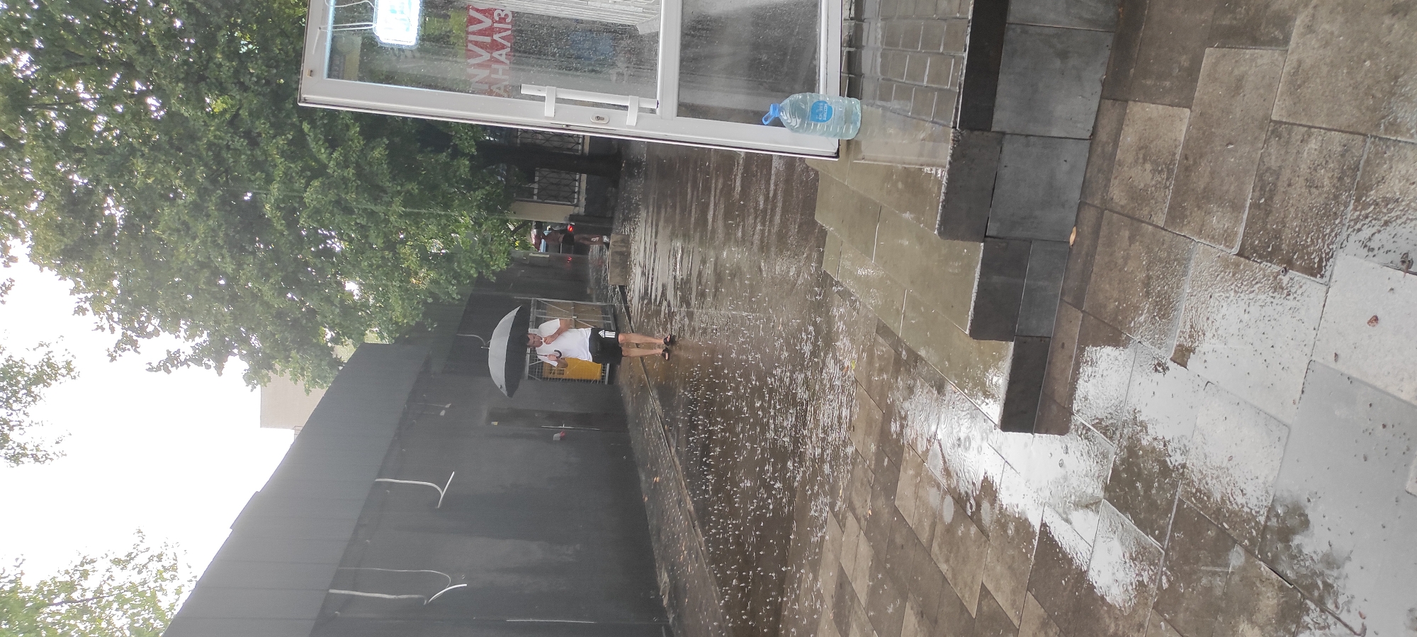 In Kamianske, Ukraine, on a rainy day, a person is walking under a black umbrella, holding it with their right hand. They are wearing a white t-shirt and black shorts with white stripes. The ground is wet and paved with square tiles, with raindrops visible on the surface. To the right of the person, there is a glass-enclosed structure with a white frame. A large blue bottle of water is placed on the steps leading up to this structure. A dark-colored building is situated to the left of the scene, with visible cables attached to its facade. Trees with green foliage are present in the background. The sky is overcast and bright.