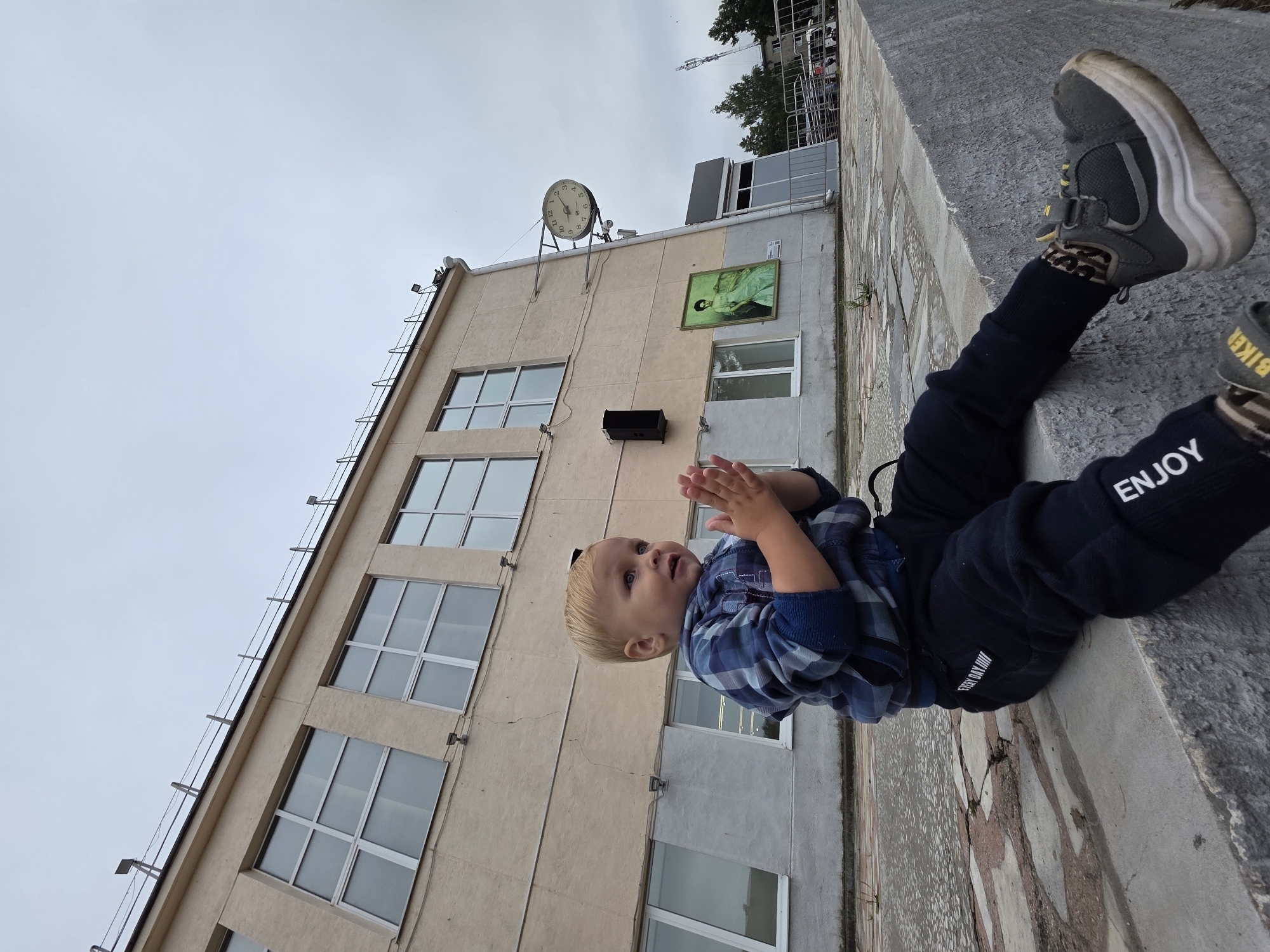 The image depicts a young boy with blonde hair and blue eyes sitting on a concrete ledge outdoors. He is wearing a blue plaid shirt, dark pants with "ENJOY" printed on one leg, and dark sneakers with yellow accents. The boy is looking to his right with his hands clasped together. Behind him is a light-colored, multi-story building with large windows. A round clock is mounted on the building's exterior, and a framed picture hangs below it. The ground in front of the building appears to be paved with cracked tiles. The sky is overcast. The scene is likely located in Chaikovsky, Russia, given the context of the surrounding information.