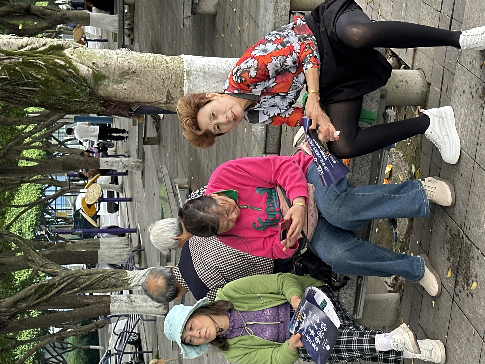 The image captures a candid outdoor scene with a group of older women sitting on a stone structure, possibly a low wall or bench. At the top of the image, a woman wearing a red and white floral shirt, black skirt, and black tights is turned toward the camera. To the left of her and below in the image, another woman, wearing a pink sweatshirt and jeans, is looking at a cell phone. Further down the frame are two more people, one in a green jacket and a hat and the other one with black-and-white patterned clothing. They appear to be reading or examining papers, possibly pamphlets or flyers. The background includes trees, benches, and other people in the distance. A sign with Chinese characters is visible. The photo seems to be taken in a public space, with the overall impression being a group of friends or acquaintances relaxing or engaging in conversation. Based on the OCR text of the image, this scene is in the Jiangjin area of China.