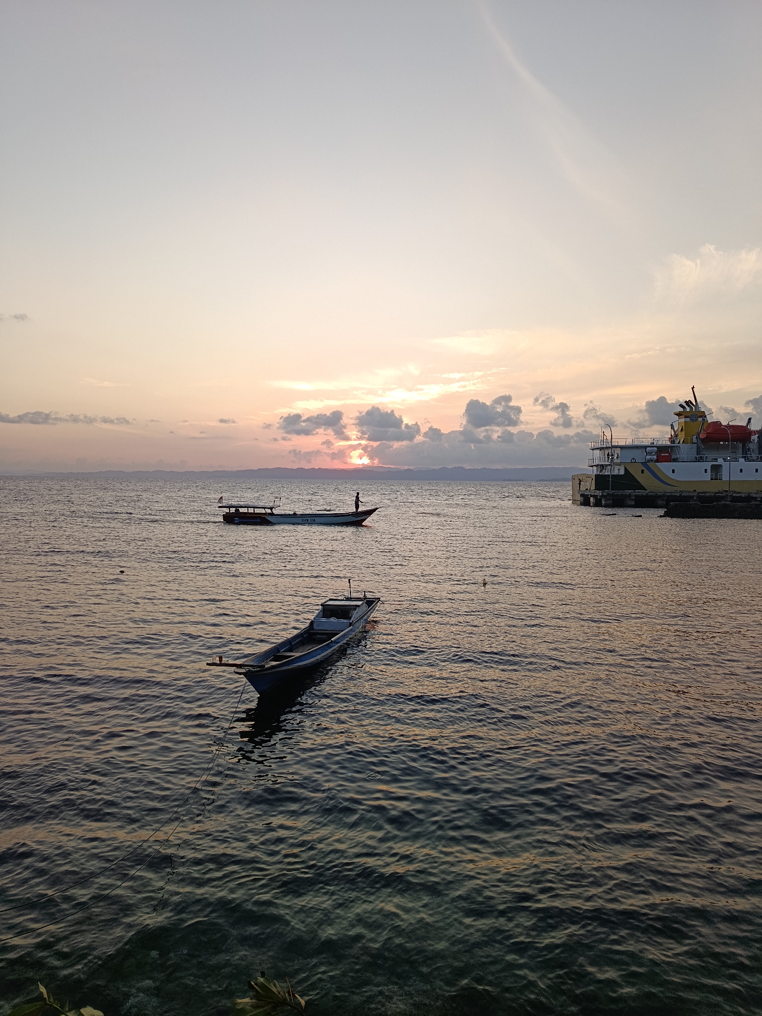 This photograph captures a tranquil scene at sunset in Mansalean, Indonesia. The sky is painted with soft hues of orange, pink, and pale blue, with scattered clouds diffusing the light. The calm sea reflects the sky's colors, creating a peaceful atmosphere.
In the foreground, a small, blue wooden boat floats gently on the water, tethered by a dark rope. Its simple design suggests it's a local fishing or transport vessel. Further out in the middle ground, another, slightly larger white boat is in motion, with a lone figure standing at its stern. This boat appears to be a motorized one, possibly a ferry or a larger fishing boat, indicated by its cabin and a small flag.
On the right side of the image, a large, multi-colored ferry or cargo ship is docked at a pier. The ship is predominantly white with yellow and blue accents, and its structure suggests it is a significant vessel. Its presence signifies a connection to wider transportation networks.
The horizon is marked by a silhouetted mountain range, adding depth to the landscape. The overall impression is one of quiet maritime activity at the end of the day, with the natural beauty of the Indonesian coast providing a serene backdrop.
