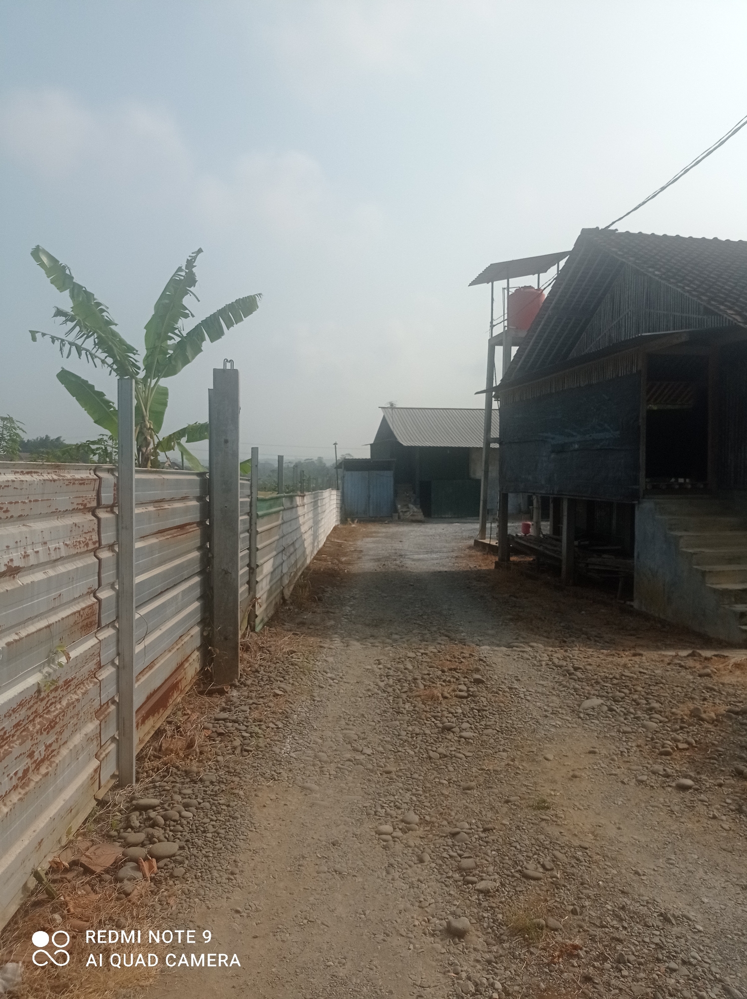 The image depicts an outdoor scene with a gravel and dirt path leading towards two buildings in Cipawon, Indonesia. To the left of the path is a corrugated metal fence, possibly a construction barrier, with visible rust and some greenery growing near its base. A tall, rectangular concrete post stands next to the fence. Further down the path, the fence extends into the distance.
On the right side of the path, a dark-colored building with a sloped, tiled roof is prominent. It appears to be elevated on stilts, with stairs leading up to an entrance area covered by a dark fabric or tarp. Adjacent to this building, to its left and further back, is another building with a similarly sloped, lighter-colored roof. A tall structure with a red water tank is visible behind and between these two buildings. In the background, a banana tree with large leaves is seen near the corrugated fence. The sky is overcast with a hazy quality. No people are visible in the image. The overall impression is of a rural or semi-industrial area.