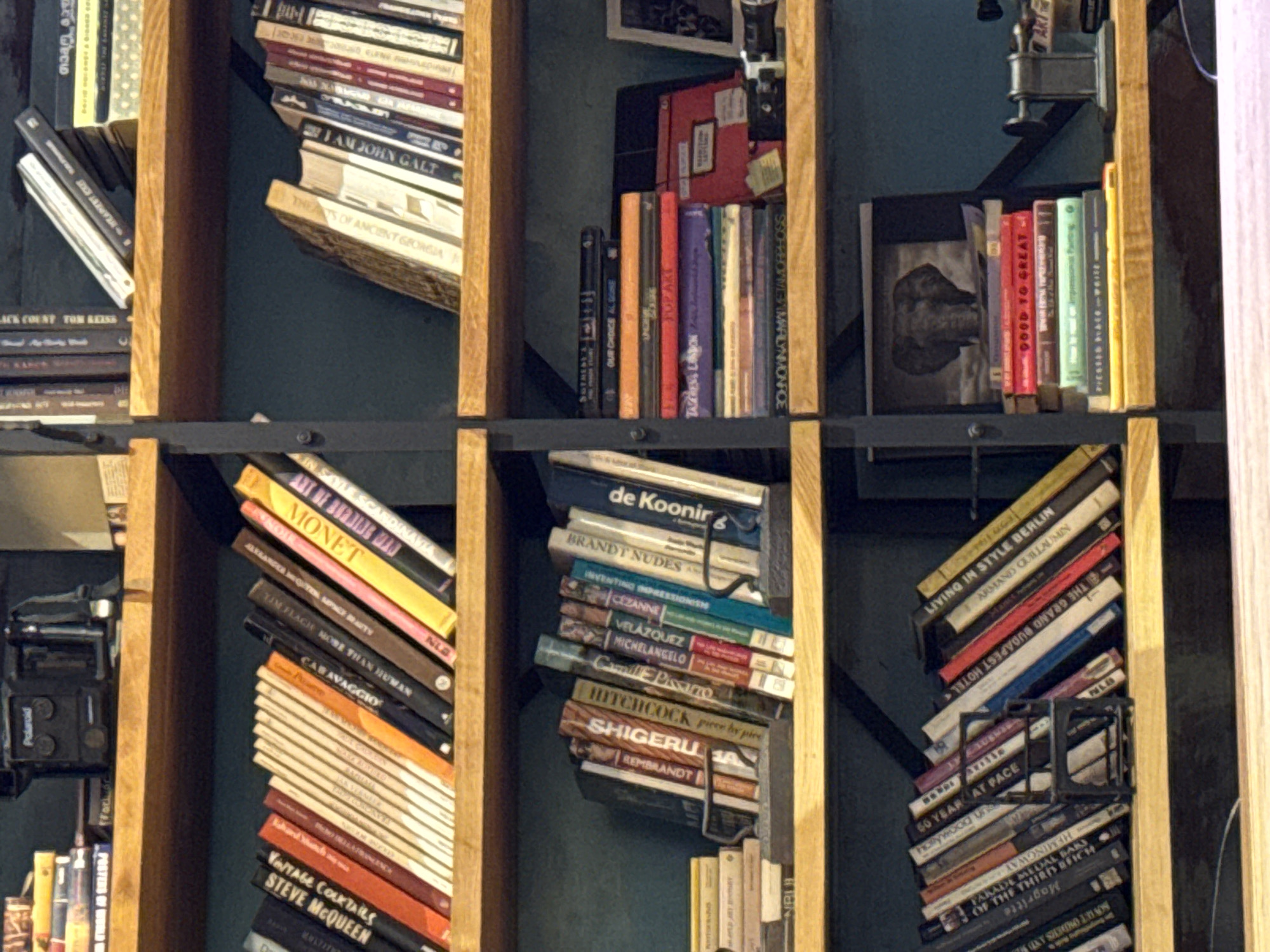 The image shows a bookshelf filled with art and literature books in a city of Tbilisi, Georgia. The books are arranged on dark metal shelves supported by a light brown wooden frame. The book titles include various subjects such as art, fashion, vintage cocktails, and biographies. The books are arranged in disarray, some leaning at angles.