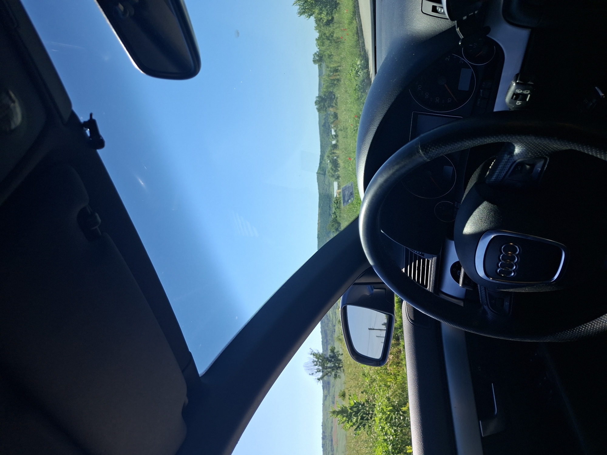 The image depicts the interior of an Audi vehicle, specifically the driver's side, with the dashboard, steering wheel, and windshield visible. Outside the windshield, a rural landscape is visible. The sky is clear and blue. The location appears to be a rural area of Yasen, Bulgaria, based on the topography, vegetation, and presence of open fields. No people are visible.