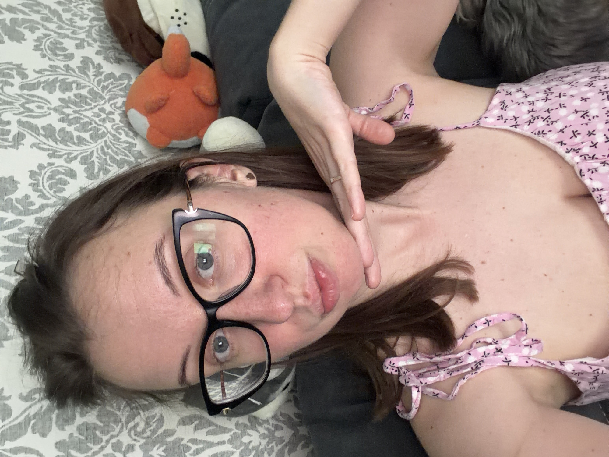 A young woman is pictured in a selfie. She is wearing large black-rimmed glasses and a pink floral-patterned top. Her right hand is resting on her chin. Behind her, a grey patterned wallpaper and stuffed animals are visible. The scene appears to be indoors, possibly in a home setting. There are no indications of this image being located in Bila Tserkva, Ukraine.