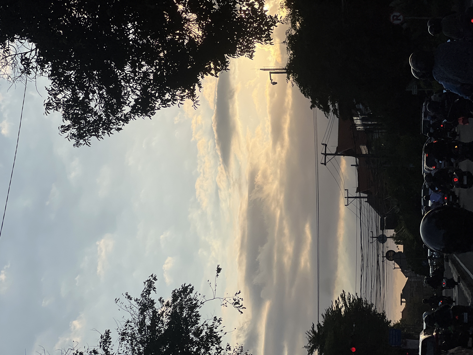 In Denpasar, Indonesia, during dusk, a busy street is depicted with numerous motorcycles and cars navigating the road. Overhead, a tangle of electrical wires crisscrosses the sky, suspended from utility poles. Silhouetted trees frame the scene, with foliage casting shadows that blend with the dimming light. The sky is a canvas of soft clouds, tinged with the warm hues of the setting sun. Buildings with traditional Indonesian architecture are visible in the background, some with terracotta-tiled roofs. A traffic light shows a red signal, indicating a temporary halt for the vehicles. The overall atmosphere is one of everyday urban life, captured as the day transitions into night.